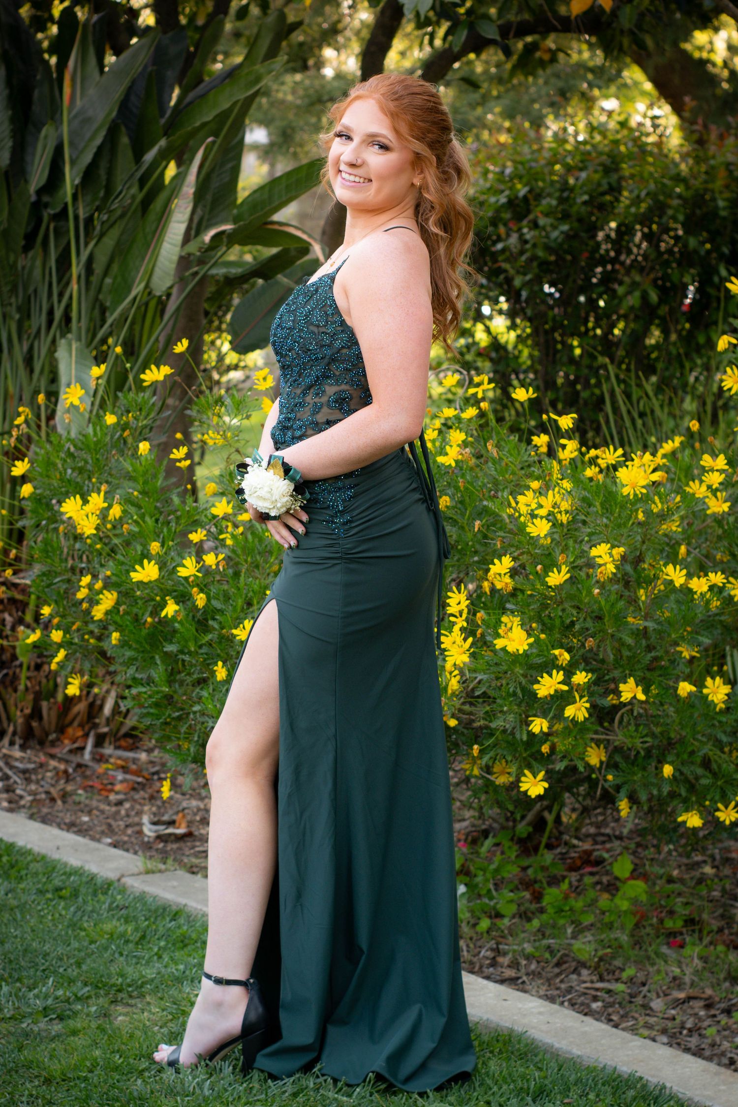 A person in an elegant emerald green dress poses beside yellow flowers in a garden setting.