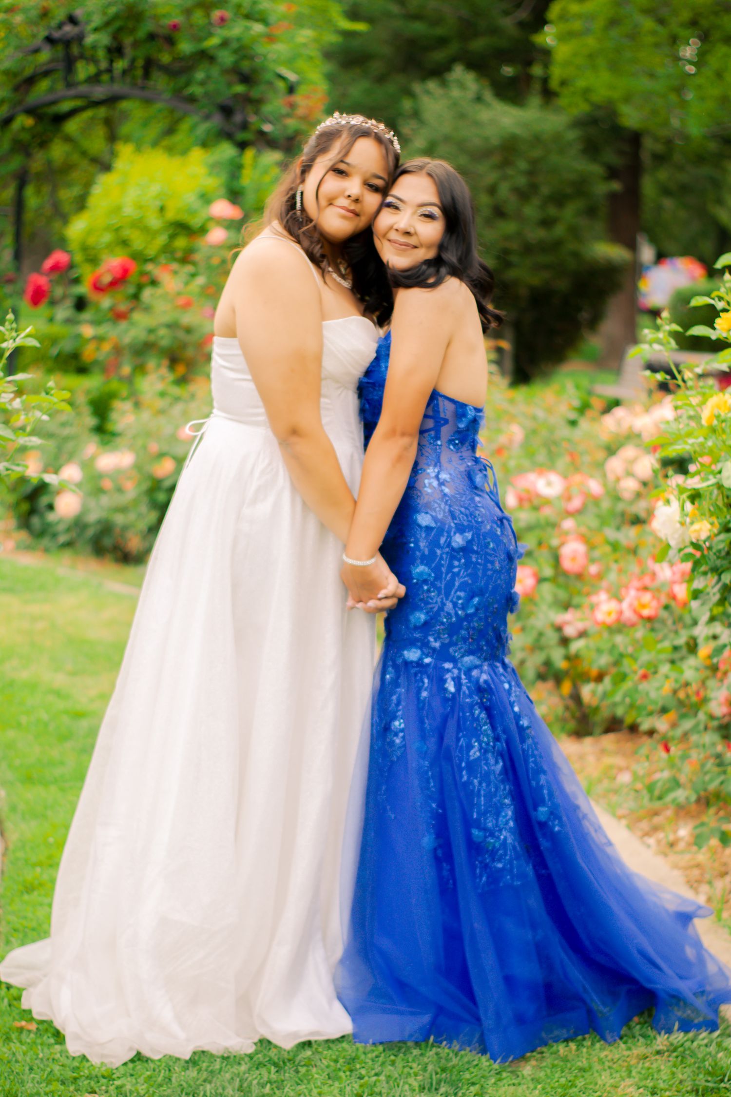 Two friends embrace while wearing formal gowns in white and royal blue against a floral garden backdrop.