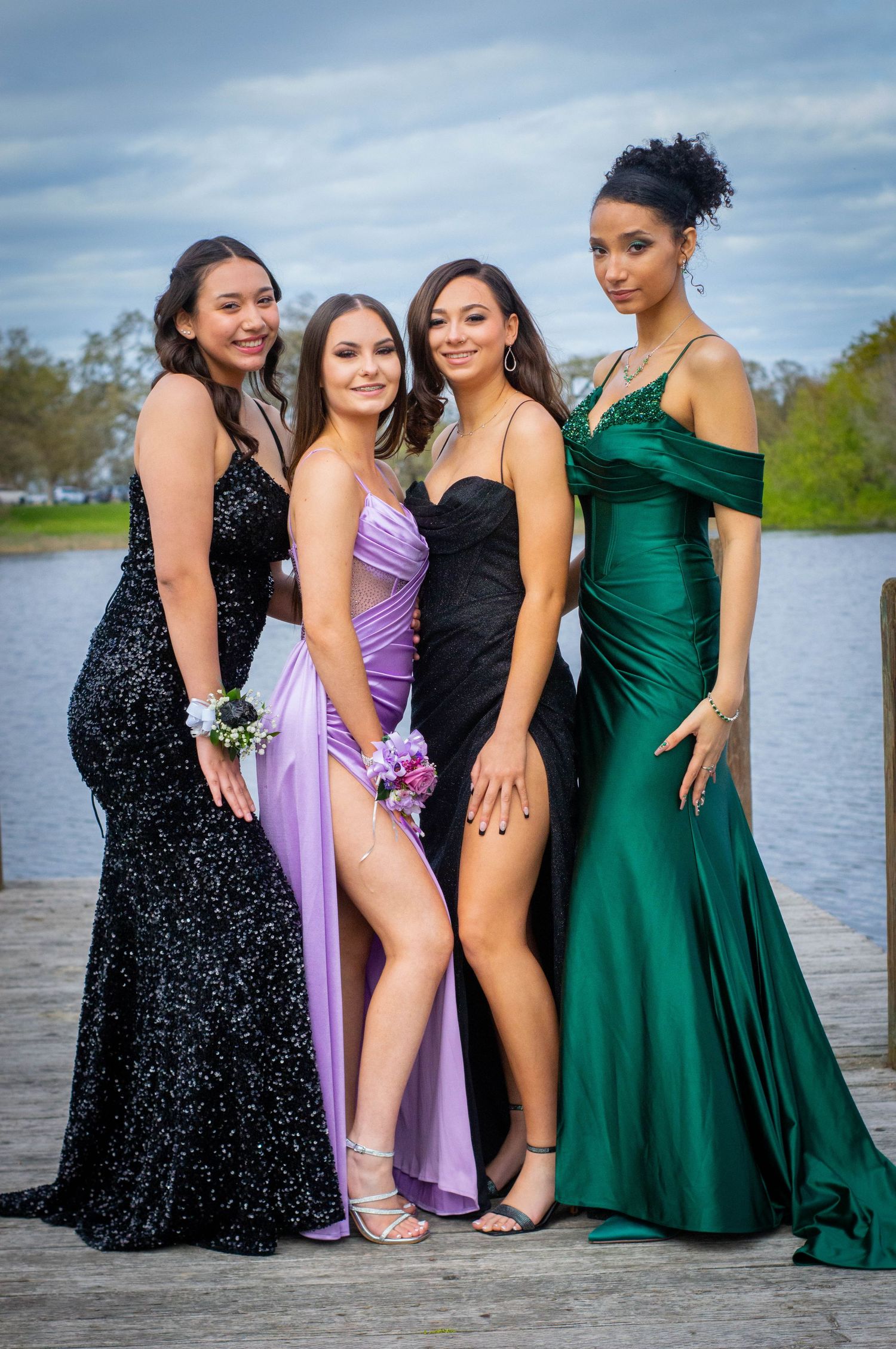 Four young women in elegant prom gowns pose on a dock, wearing corsages in black, purple, and green dresses.