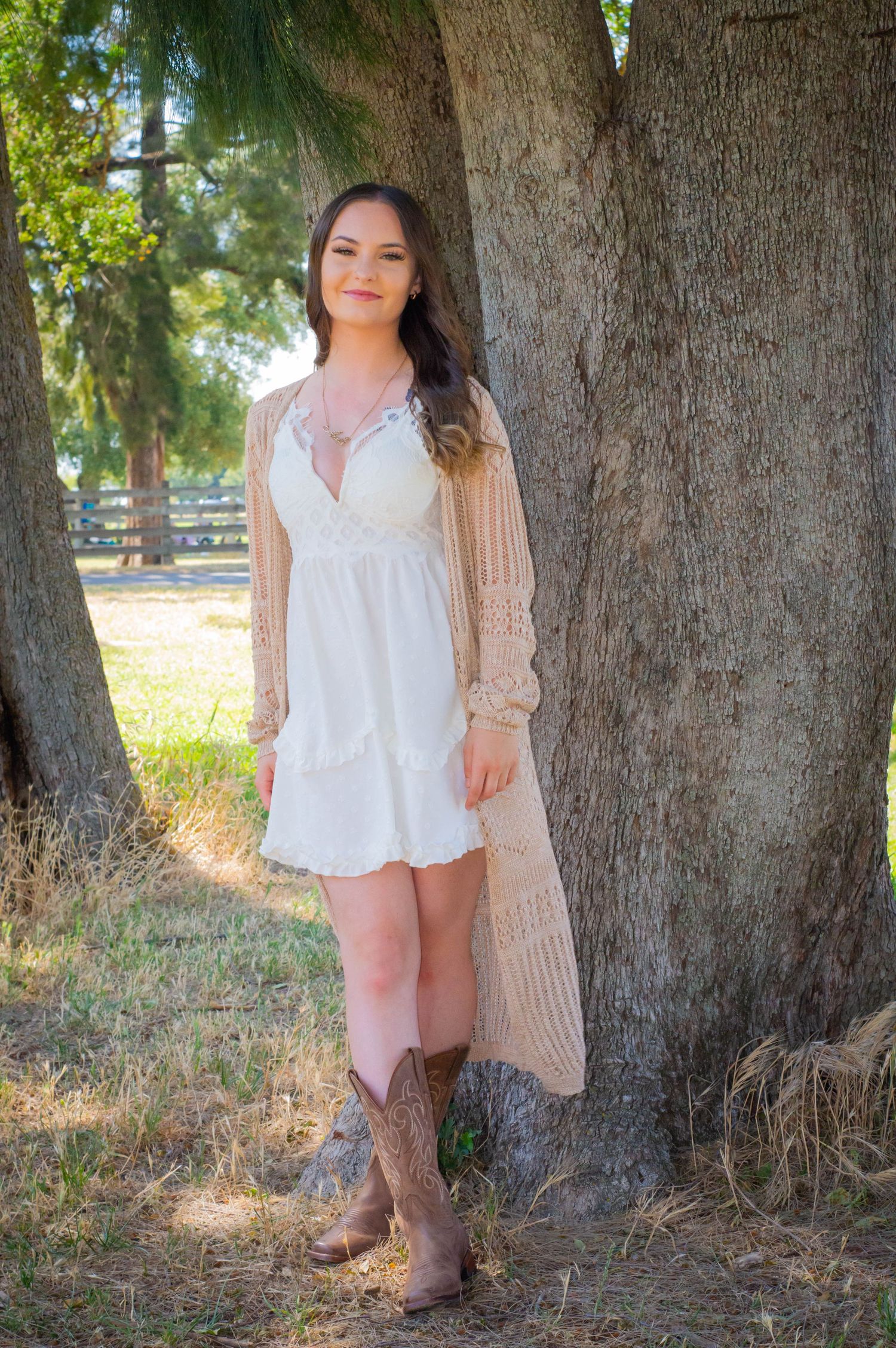 Person wearing white dress and boots poses near large tree trunk in natural outdoor setting.