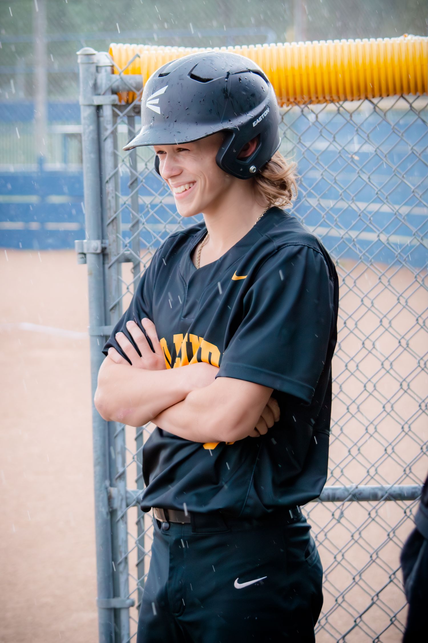 High school baseball player in black uniform standing by chain link fence during softball game.
