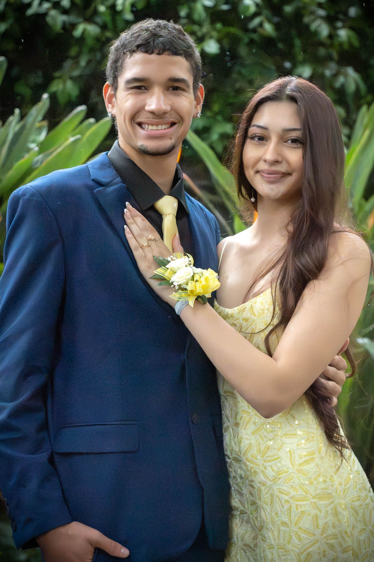 A young couple in prom outfits pose outdoors, the woman placing her corsaged hand on the man's chest.