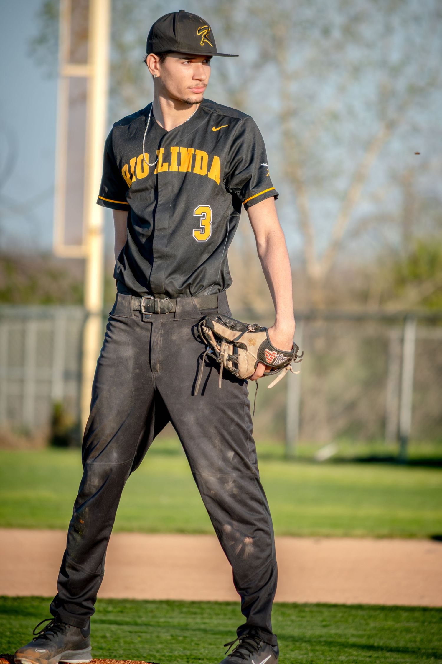 Baseball player #3 in a dark Yorba Linda uniform stands on the field holding a glove, pants dirty from play.