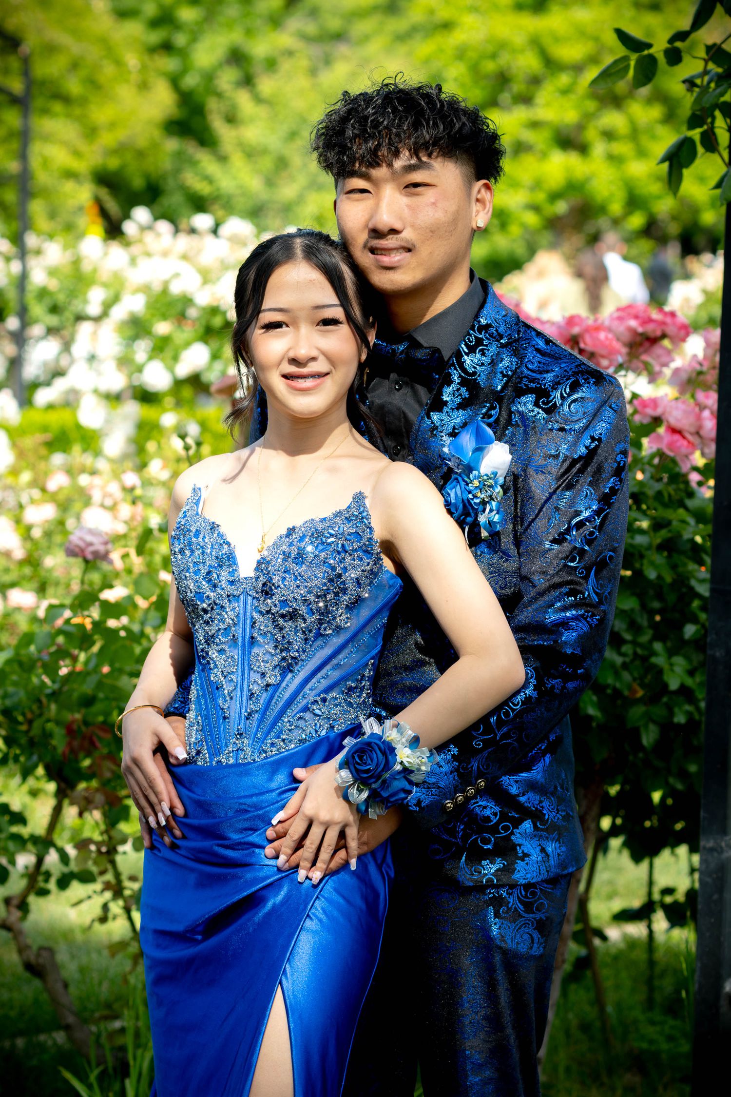 Prom couple in matching blue outfits pose together in rose garden.