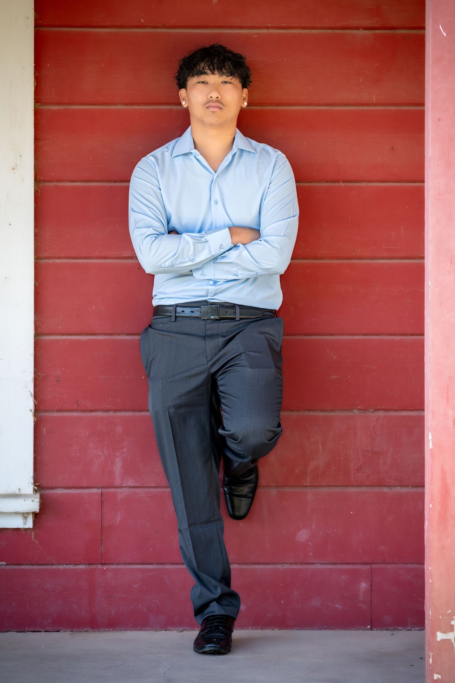 Young man in blue dress shirt and gray slacks leaning against red wall with arms crossed.
