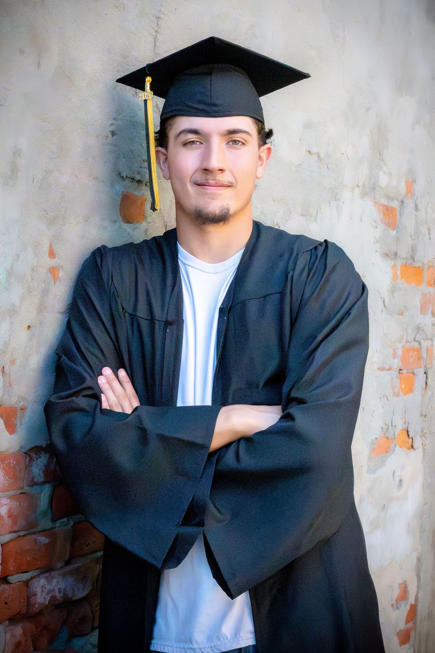 Young man in black graduation cap and gown stands confidently with arms crossed against a rustic brick wall.