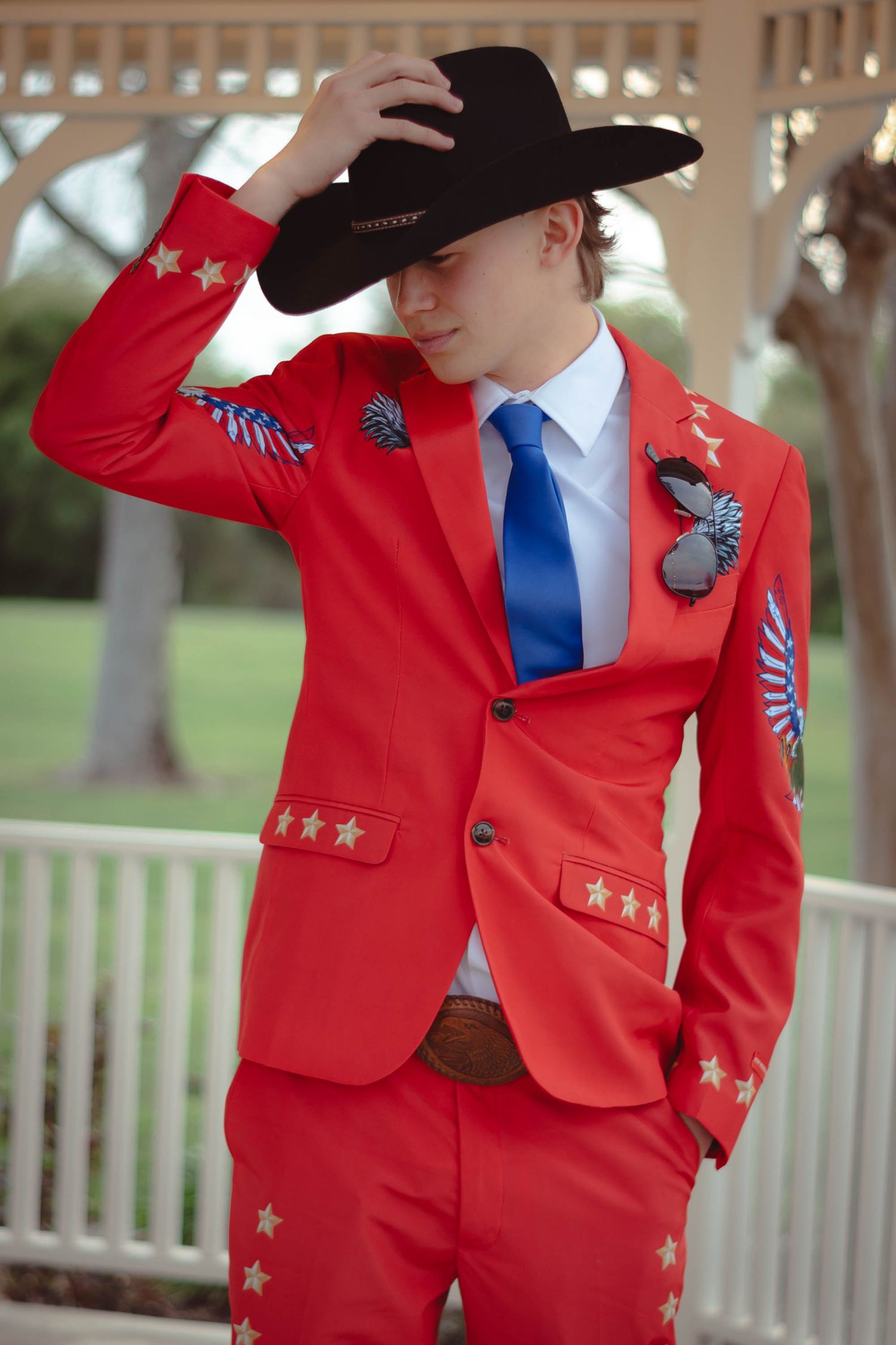 Person in red formal jacket and black cowboy hat strikes poses against white gazebo backdrop.