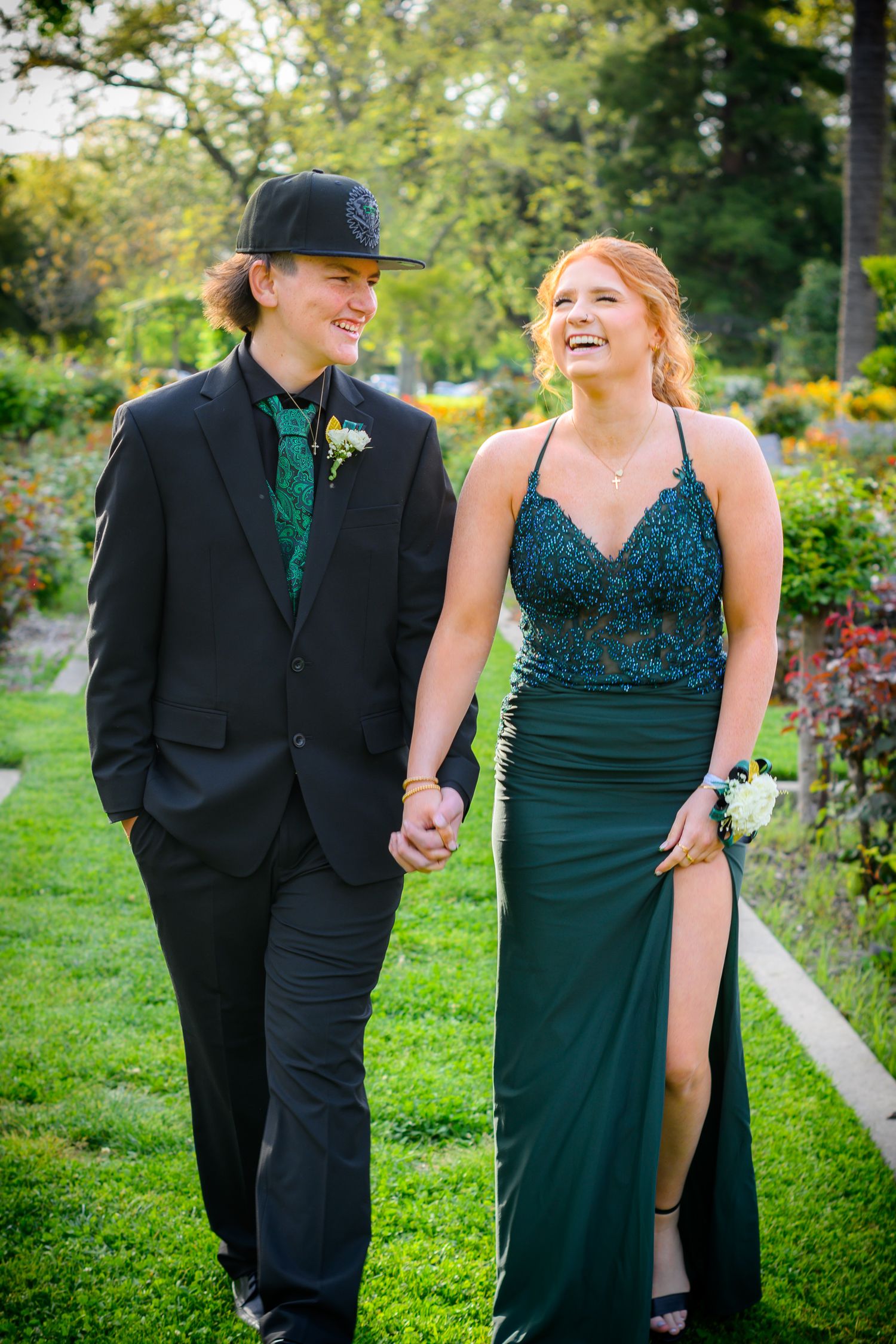 A couple walks hand in hand through a garden while wearing coordinating formal wear in black and green.