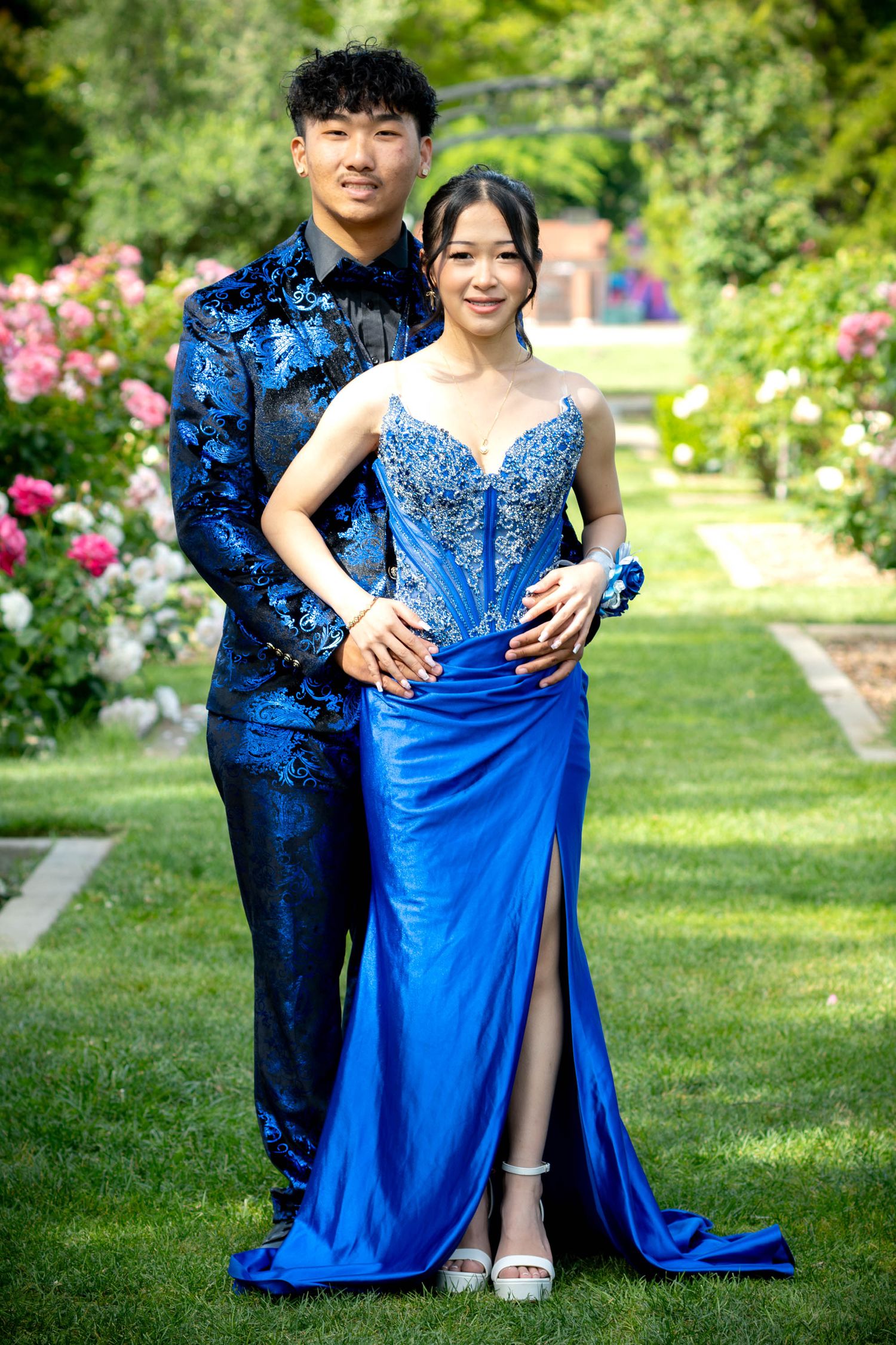 Young couple in matching royal blue prom outfits posing in a rose garden, wearing elegant formal wear.