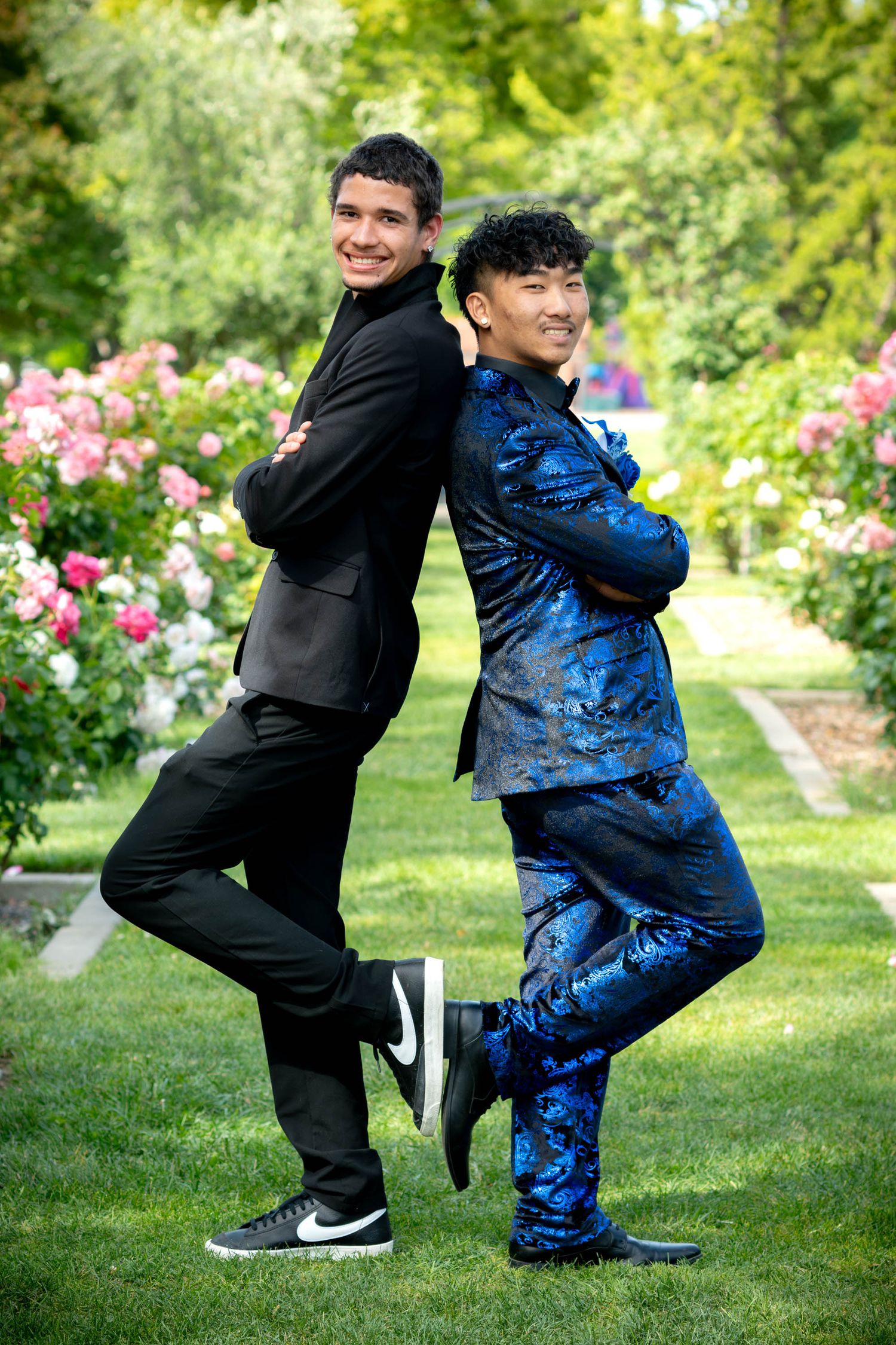 Two young men in formal suits pose back-to-back in a rose garden, one in black and one in a blue patterned suit.