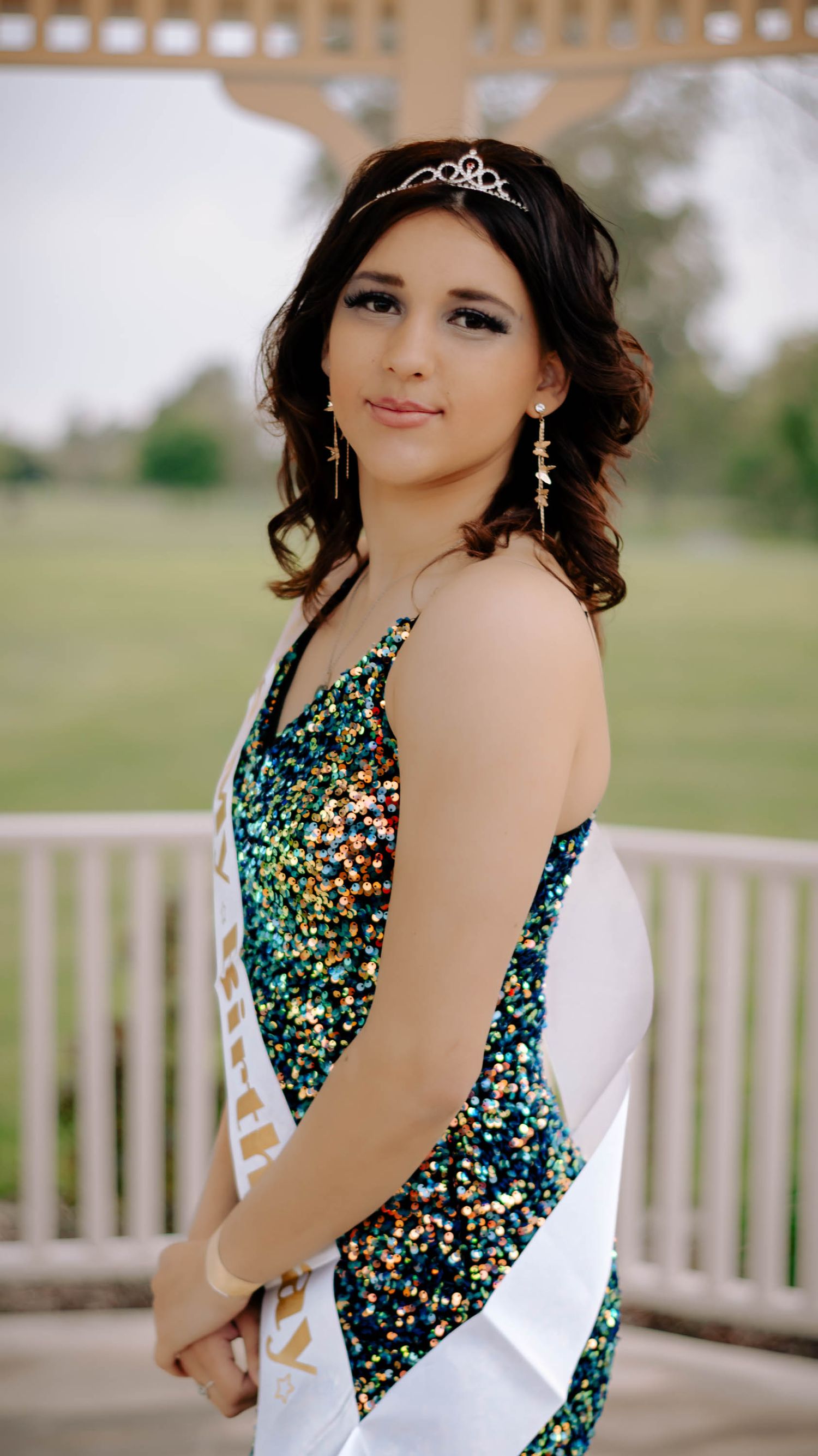 Person in sparkly formal dress with tiara poses on white gazebo platform.