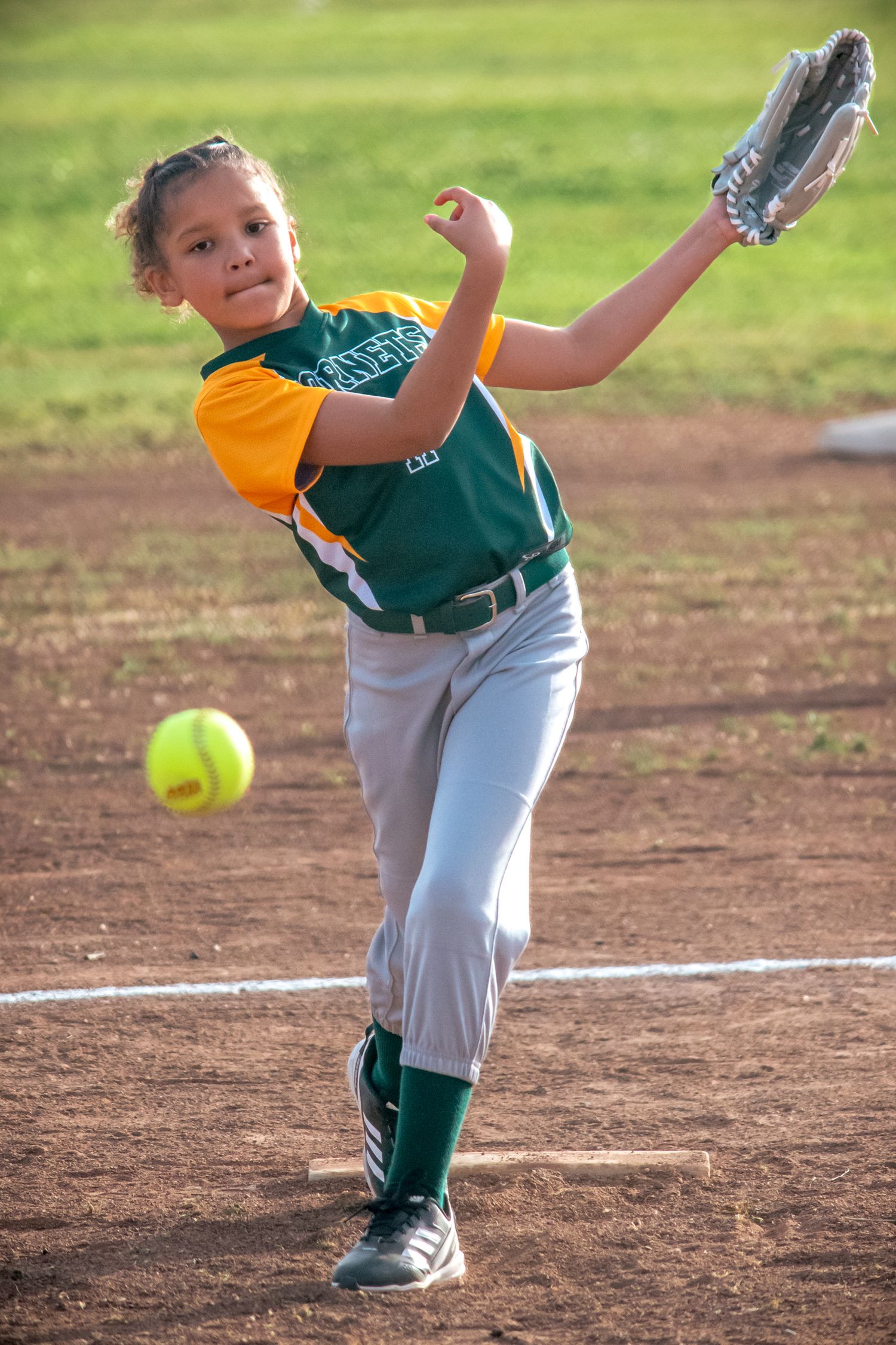 Young pitcher in green uniform executing a pitch delivery during a softball game.