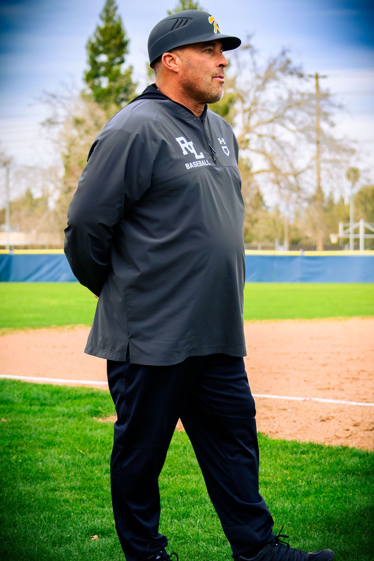 Baseball coach in black uniform walking on baseball field during practice.