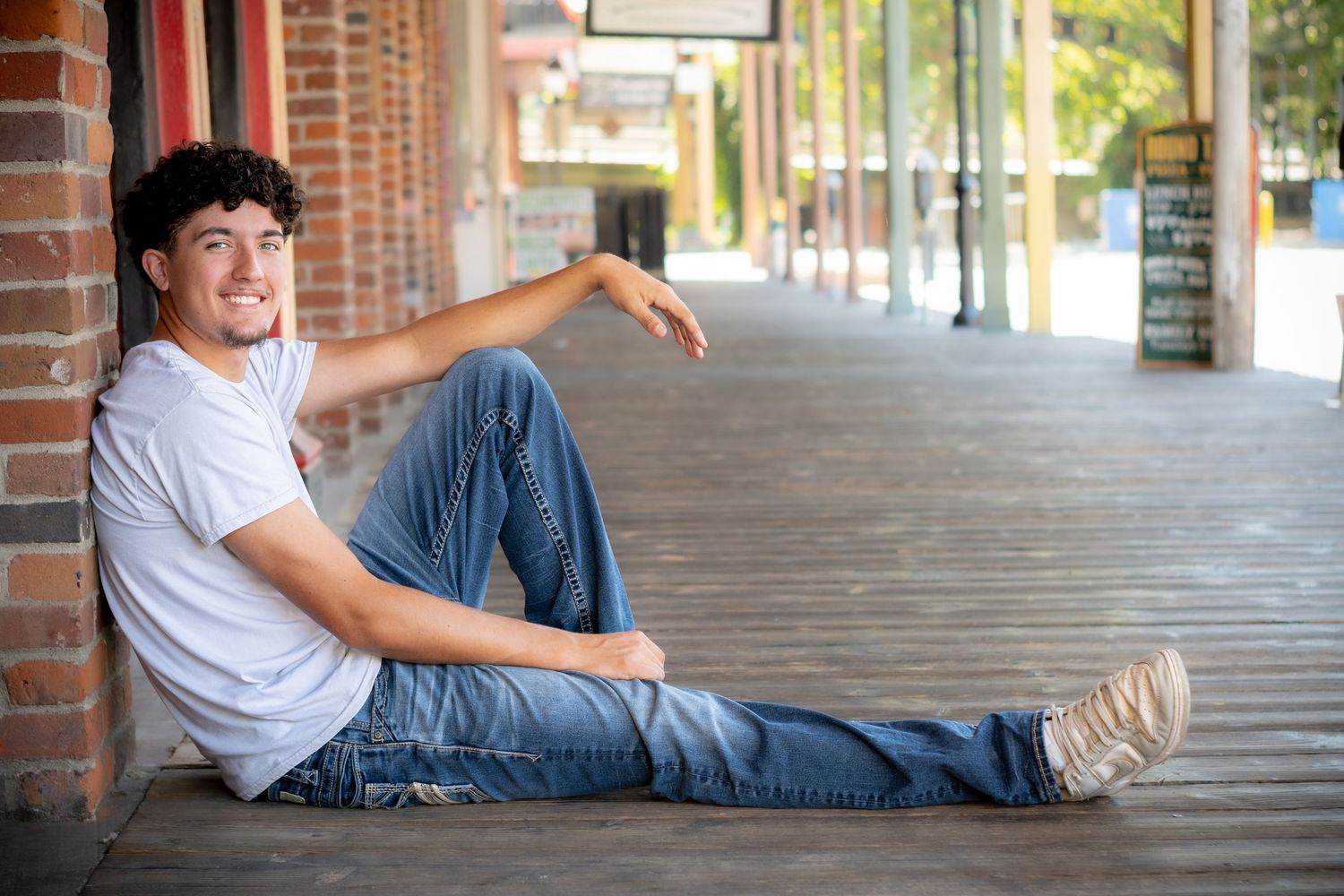 Young man in white t-shirt and jeans sitting relaxed on a wooden sidewalk, smiling confidently outdoors.