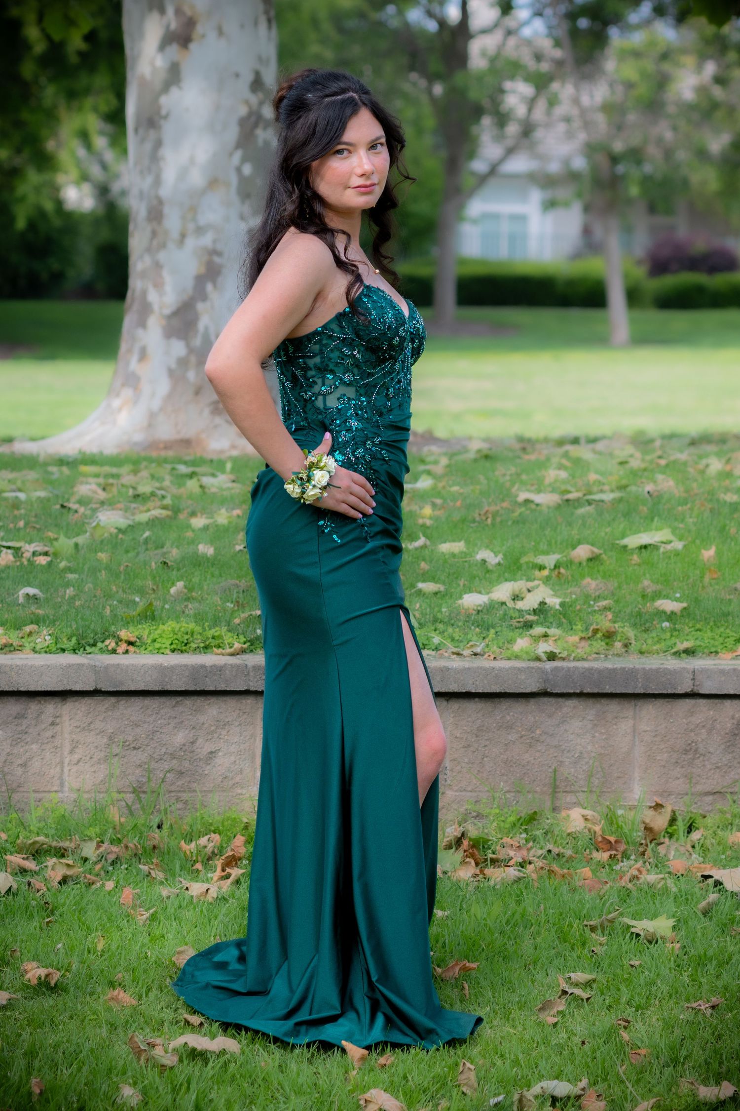 Young woman in elegant emerald green sequin prom gown with slit and wrist corsage poses in a park setting.
