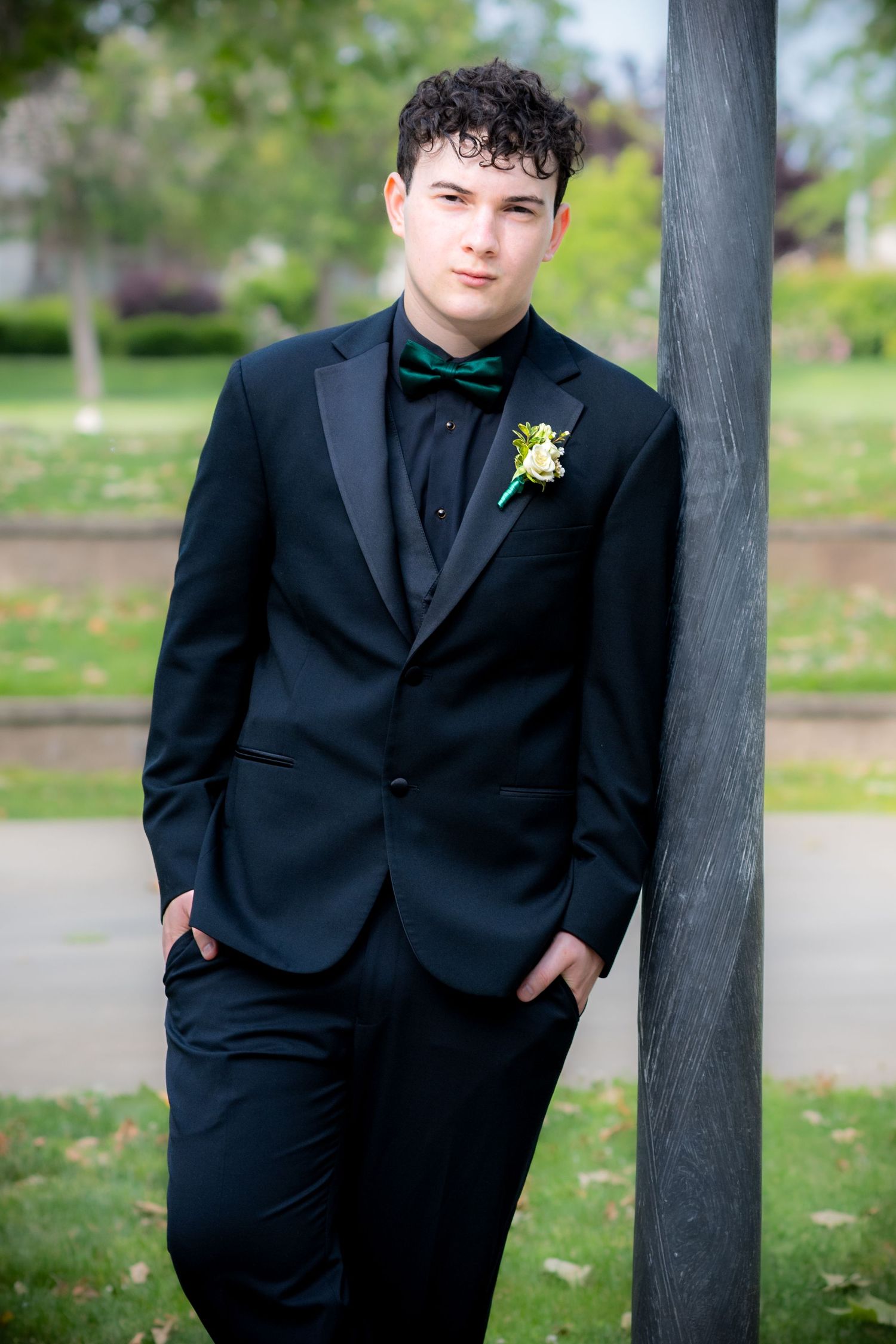Young man in black tuxedo with green bow tie and boutonniere leaning against a pole outdoors.