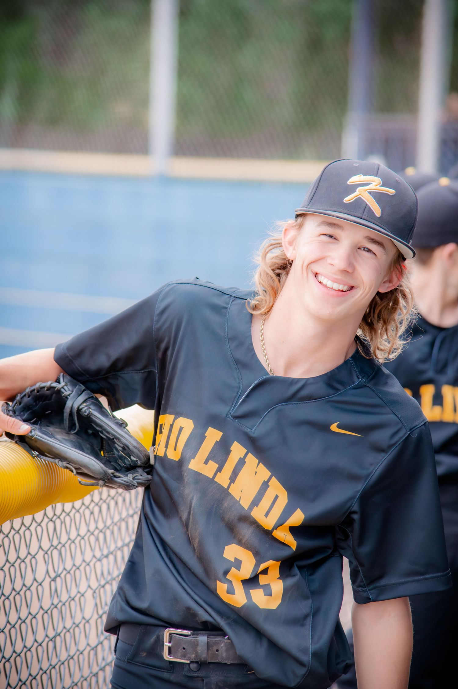 Baseball player in black uniform jersey posing against chain link fence.
