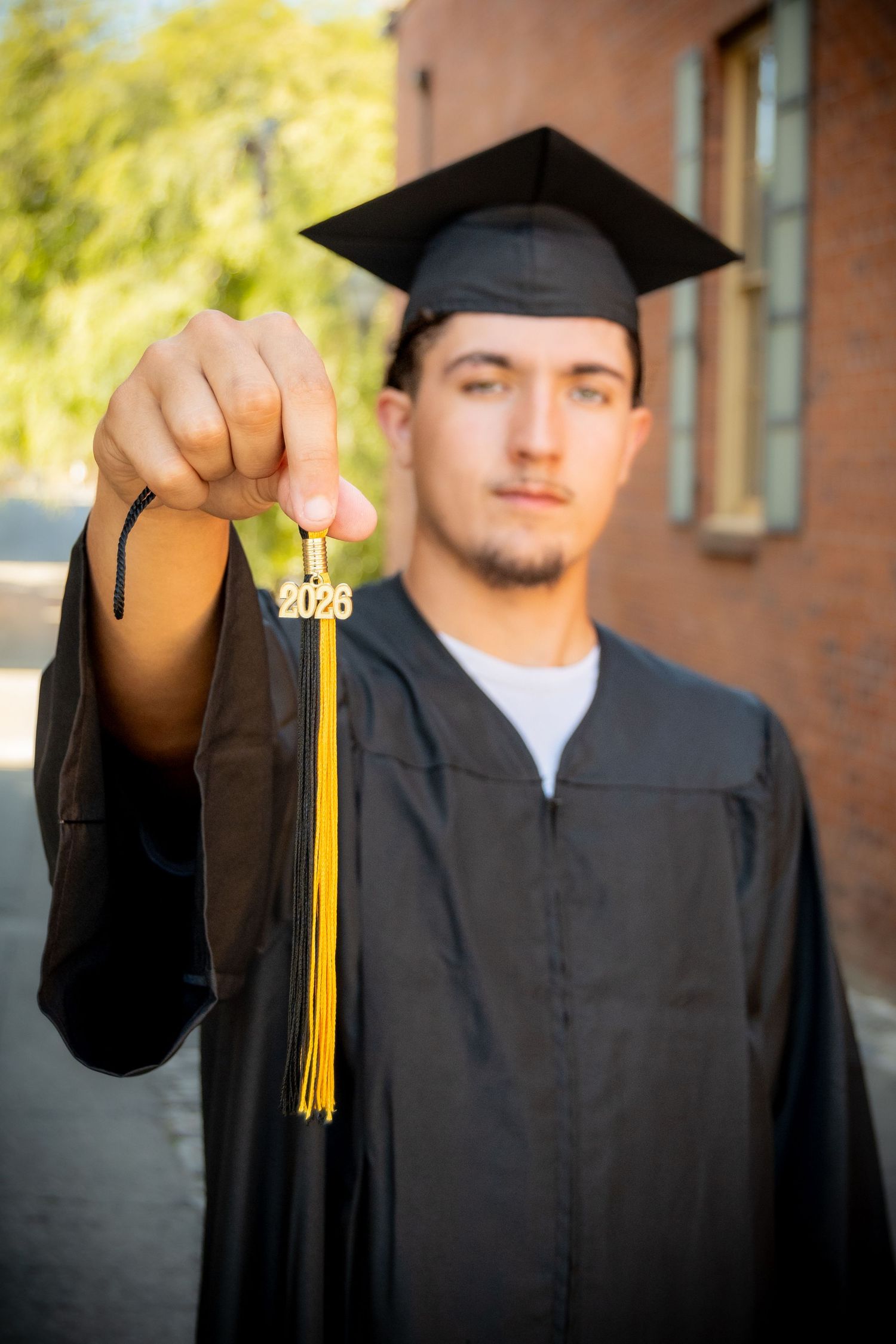 Graduate in black cap and gown holds yellow 2023 tassel toward camera outdoors near brick building.