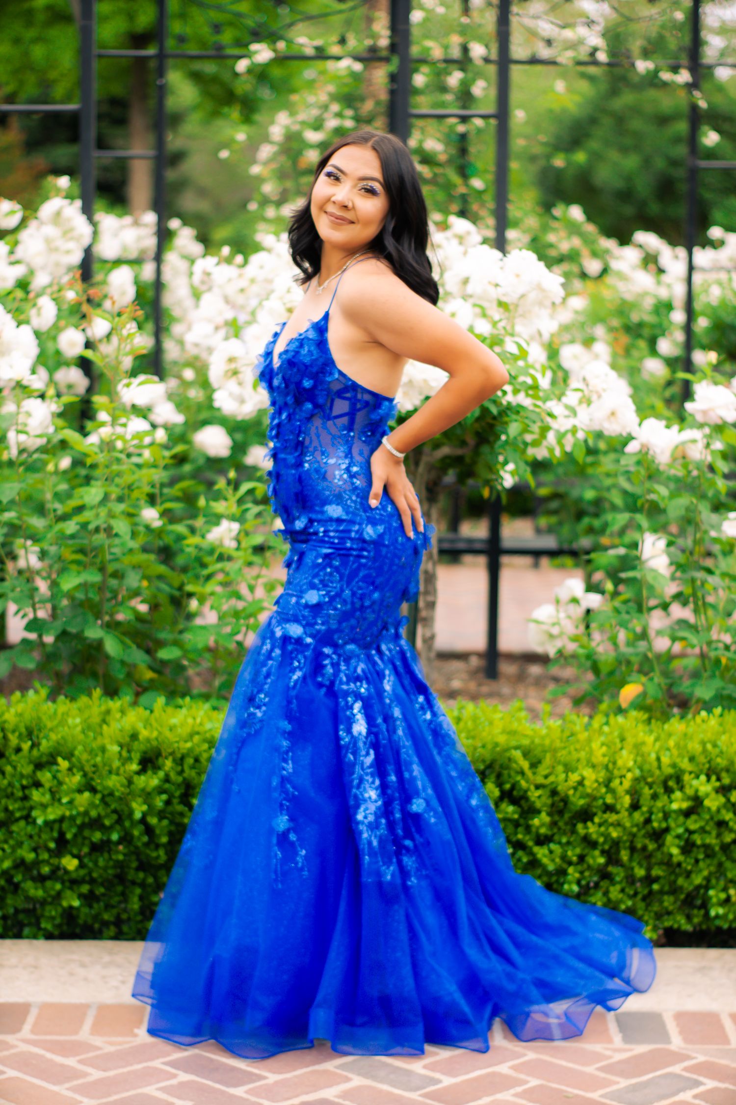 A woman in a stunning royal blue mermaid gown poses in a garden setting with blooming flowers.