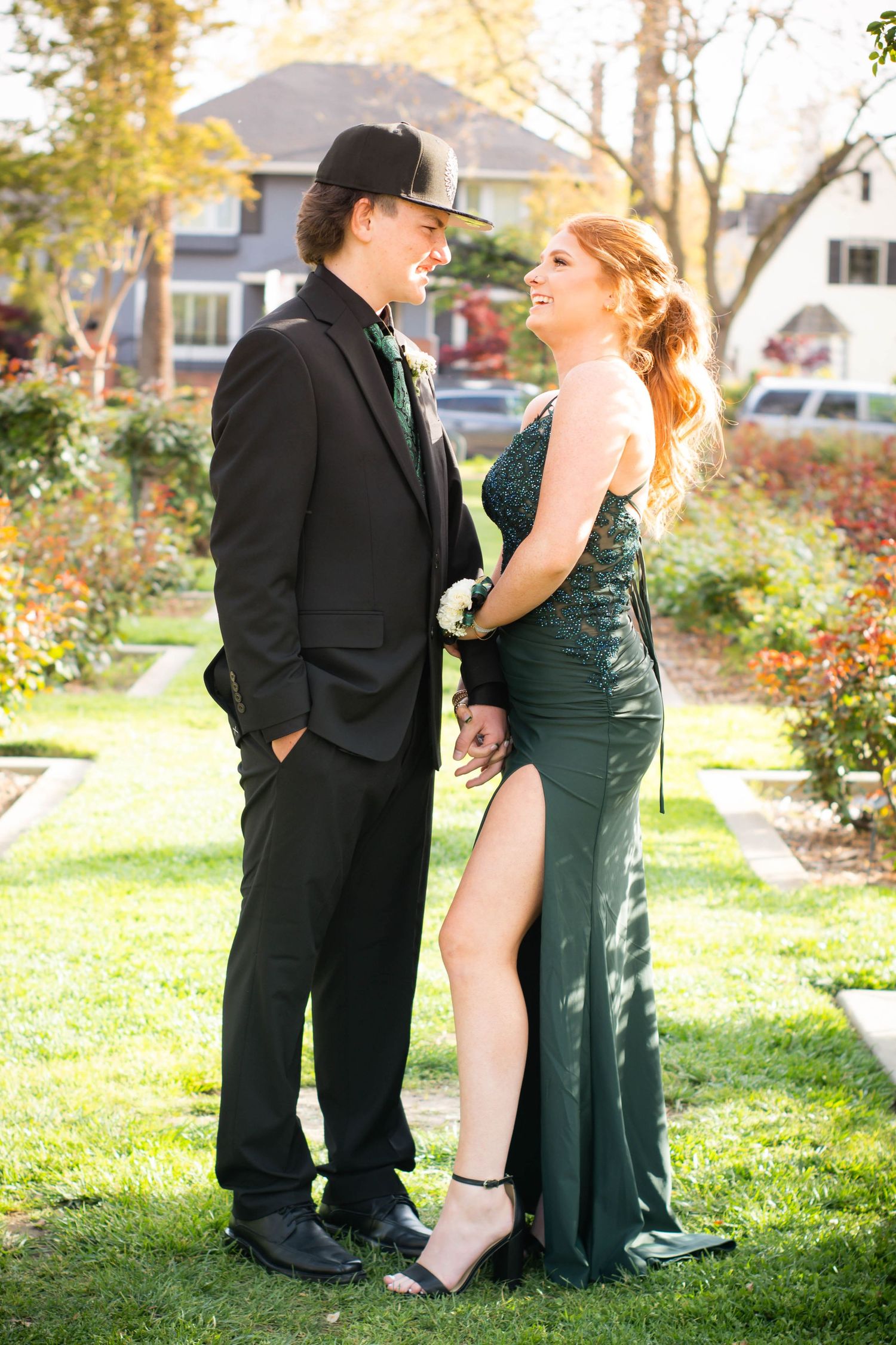 A couple in formal wear pose together in a garden, wearing black suit and emerald green dress.