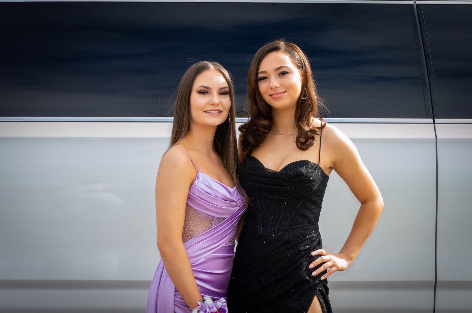 Two young women in elegant prom dresses, one in purple and one in black, posing together outdoors.
