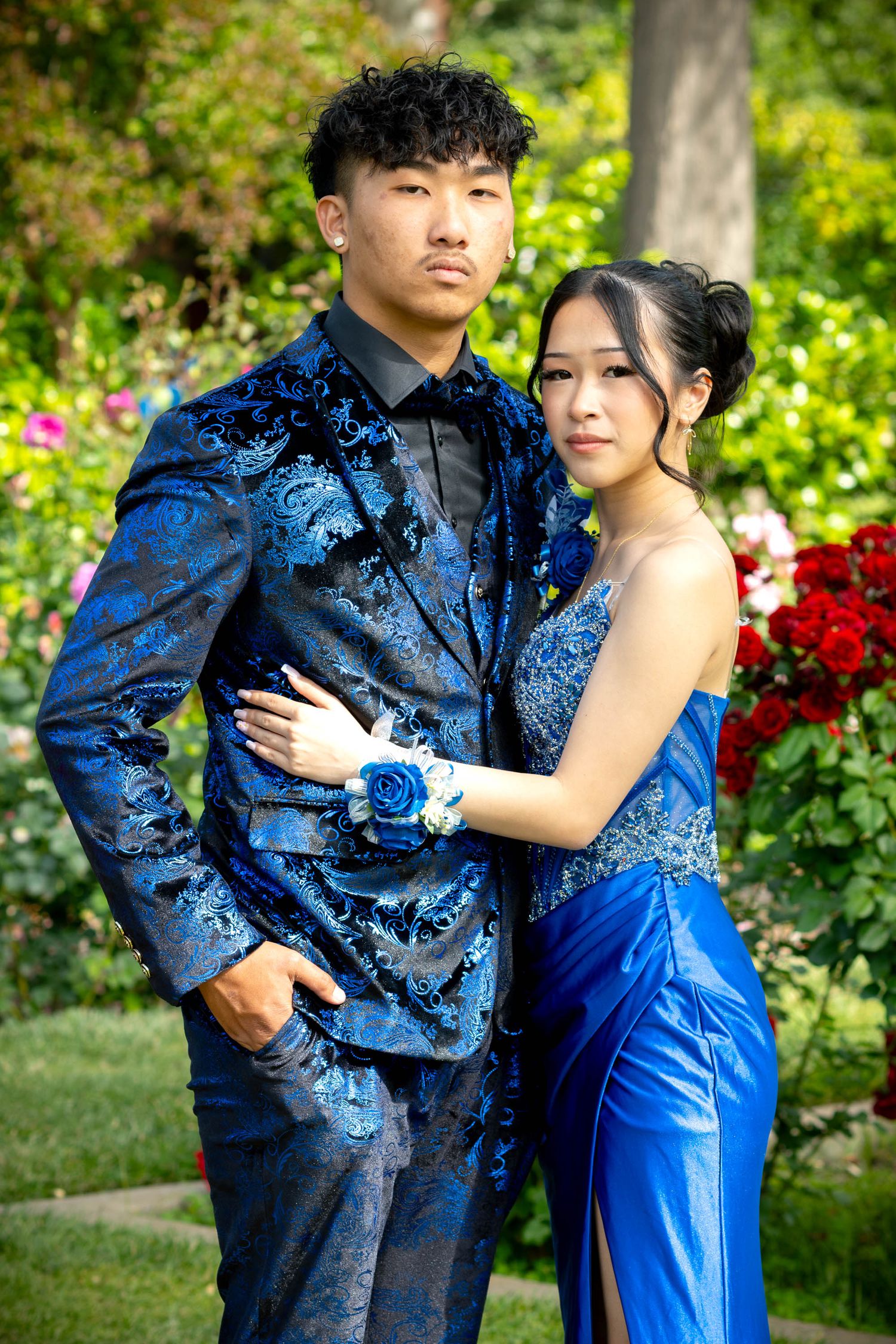 A young Asian couple in matching royal blue prom outfits pose in a garden with red roses.