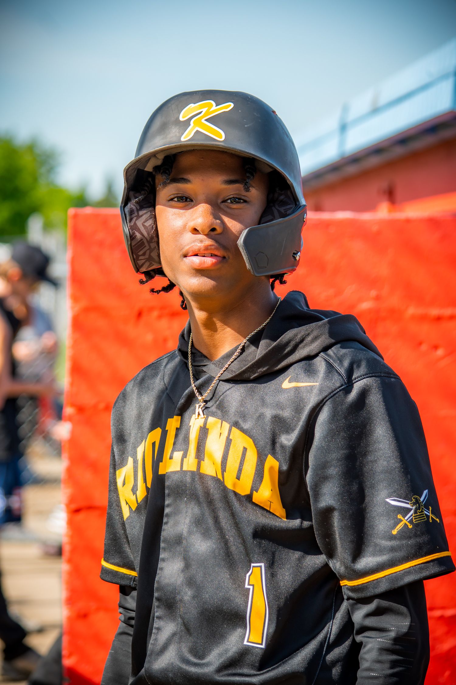 Young baseball player in black Richland uniform with number 1 jersey and batting helmet poses against red background.