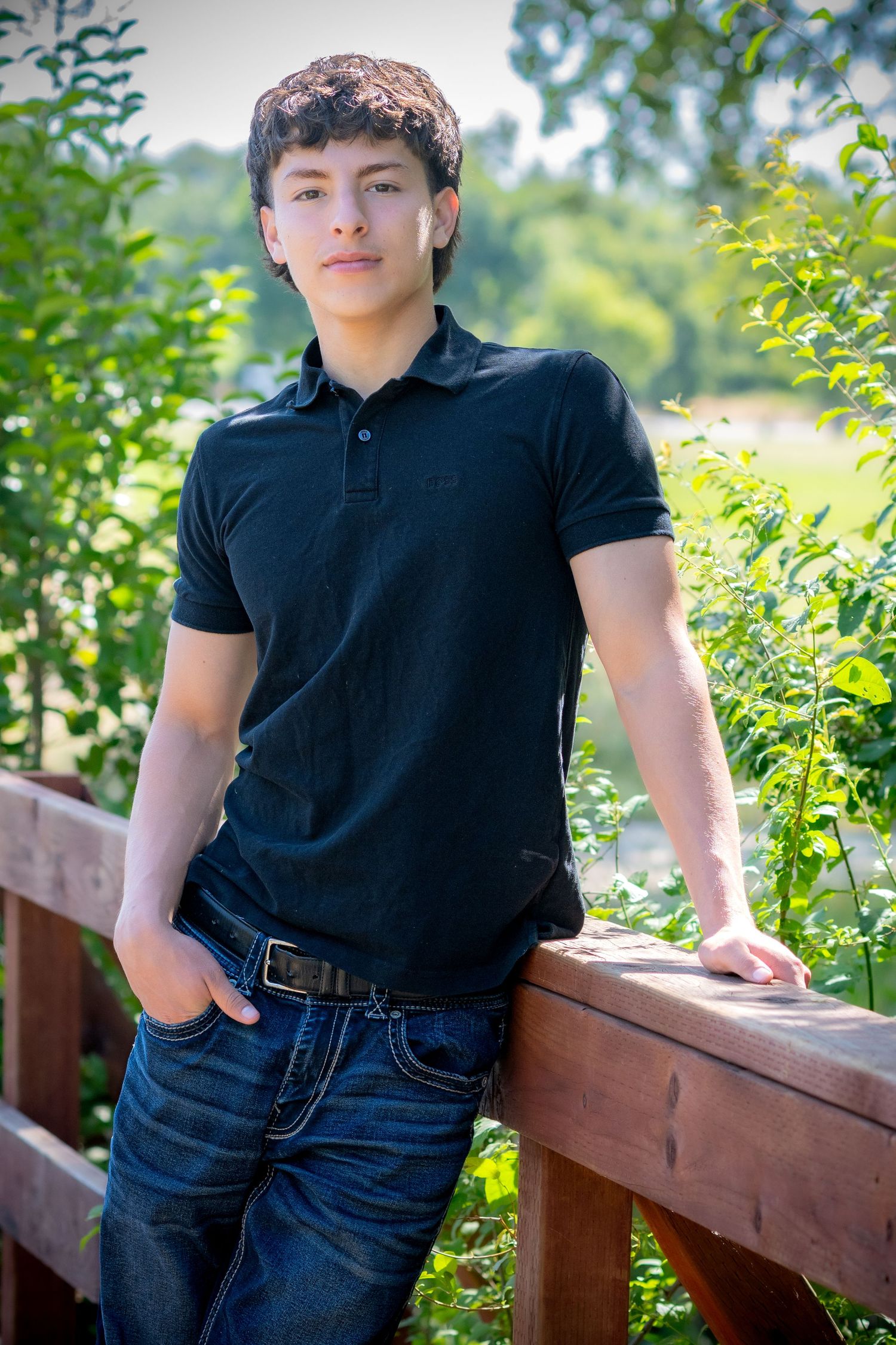 Teen boy in black polo and jeans leans on wooden railing surrounded by lush green foliage outdoors.