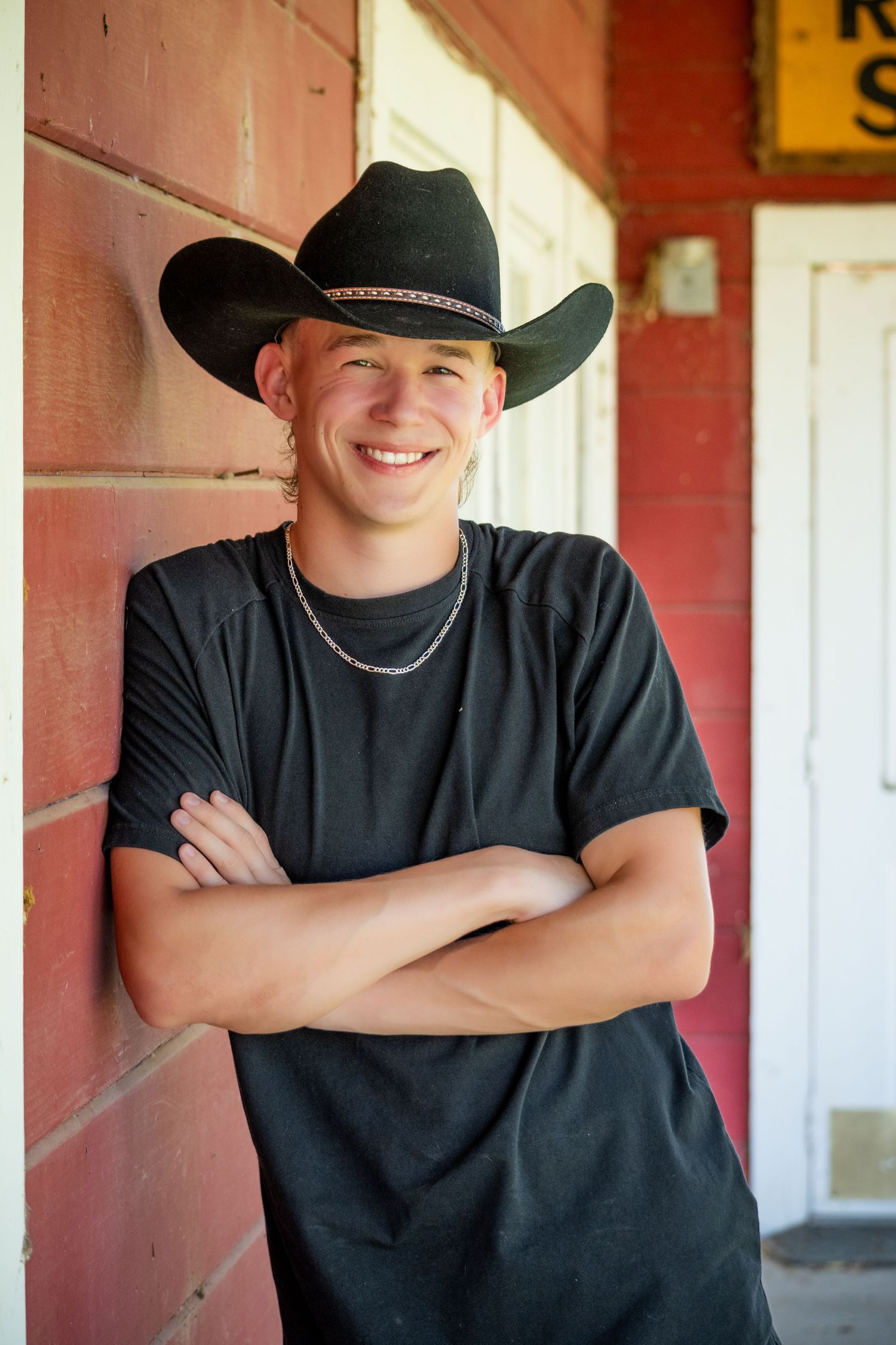 Western style portrait session against red barn wall wearing black shirt and cowboy hat.