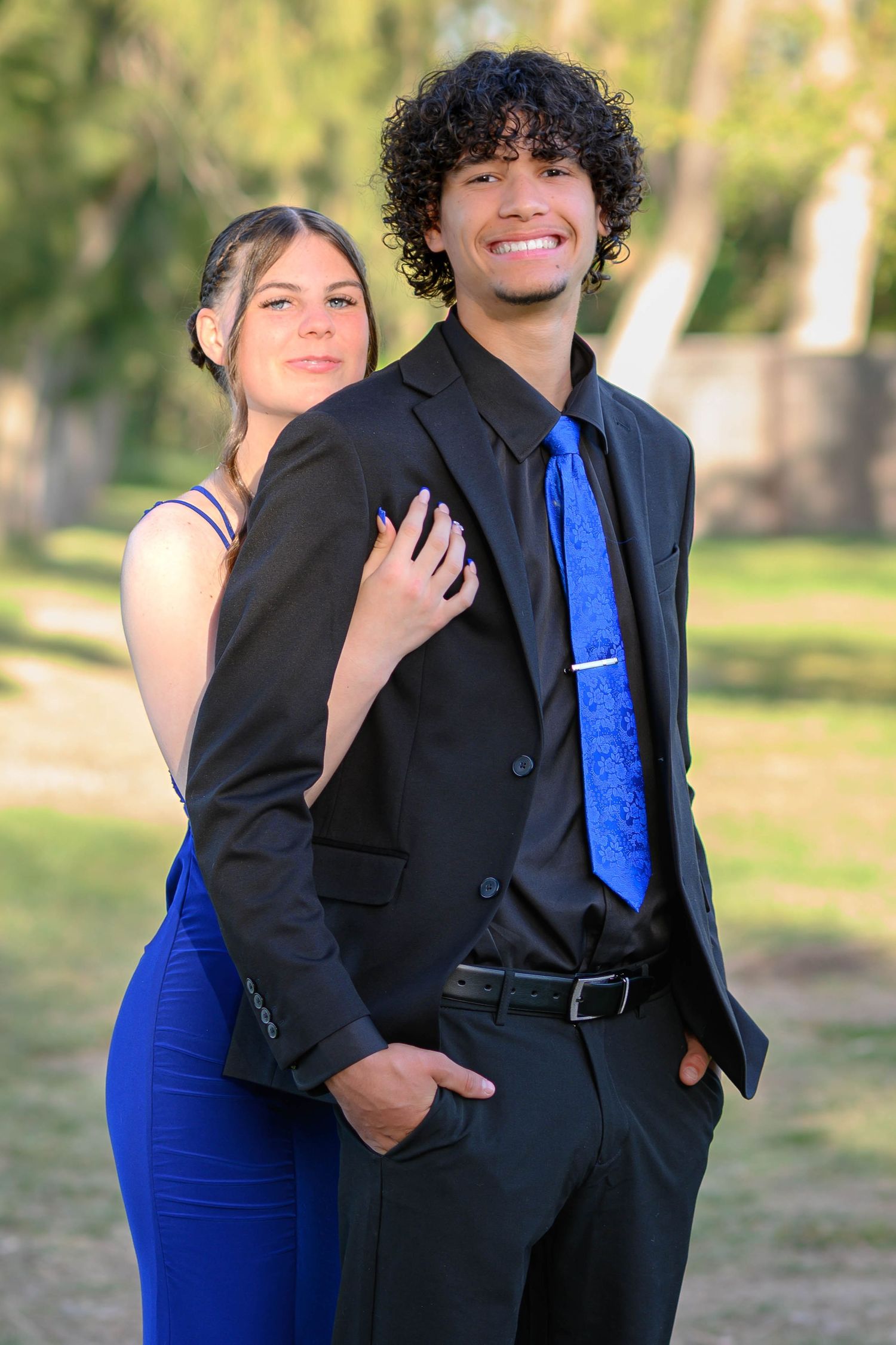 A couple in formal attire pose together outdoors, with one wearing a black suit and blue tie.