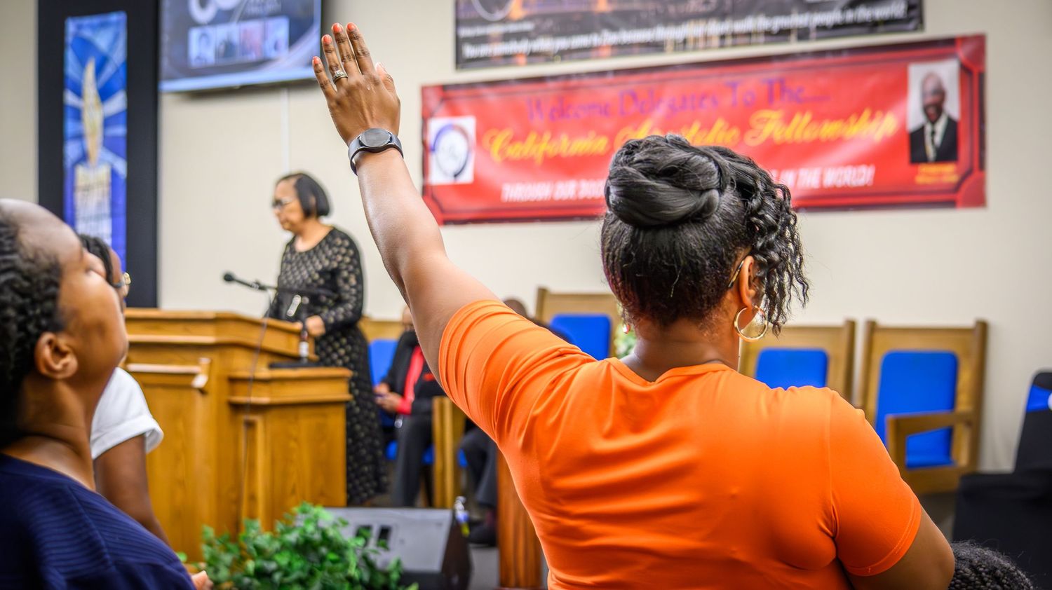 People raising hands in worship with orange and red clothing visible in an indoor church setting.