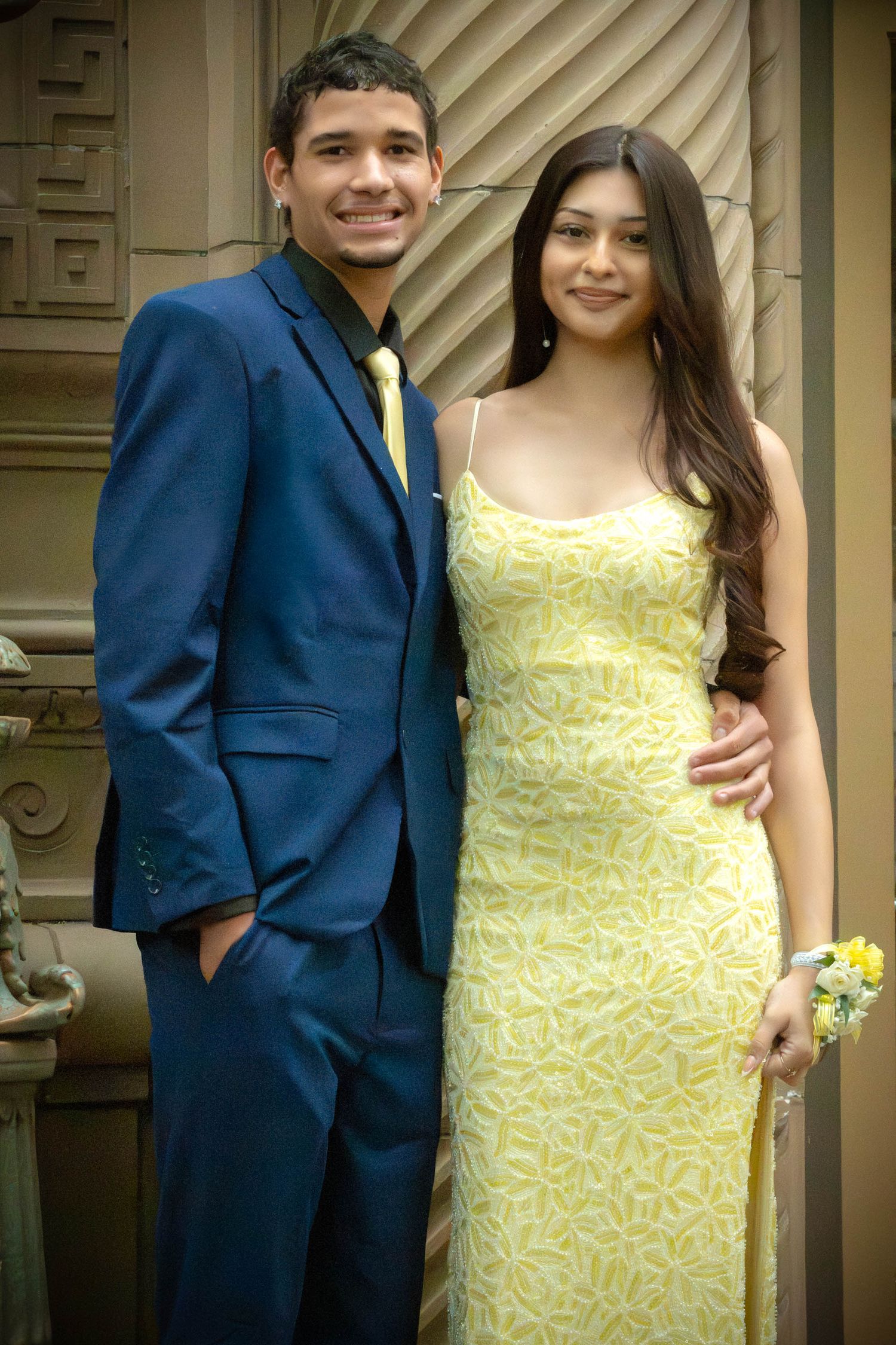 A young couple dressed for prom, man in blue suit with yellow tie, woman in yellow floral gown with wrist corsage.