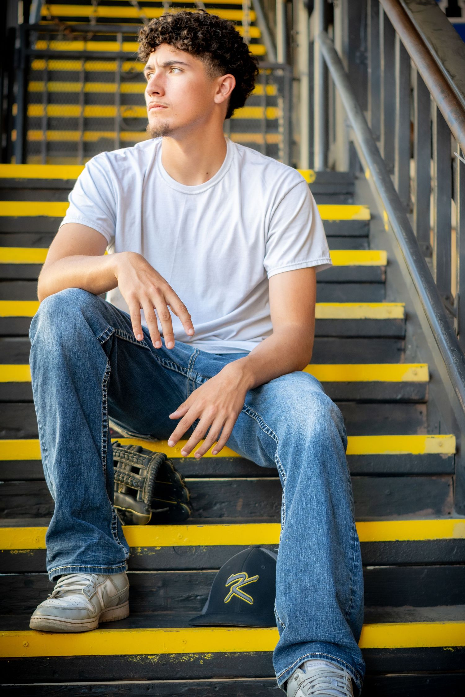 Young man with curly hair sits on yellow-striped bleacher steps wearing white tee and jeans, holding a baseball glove.