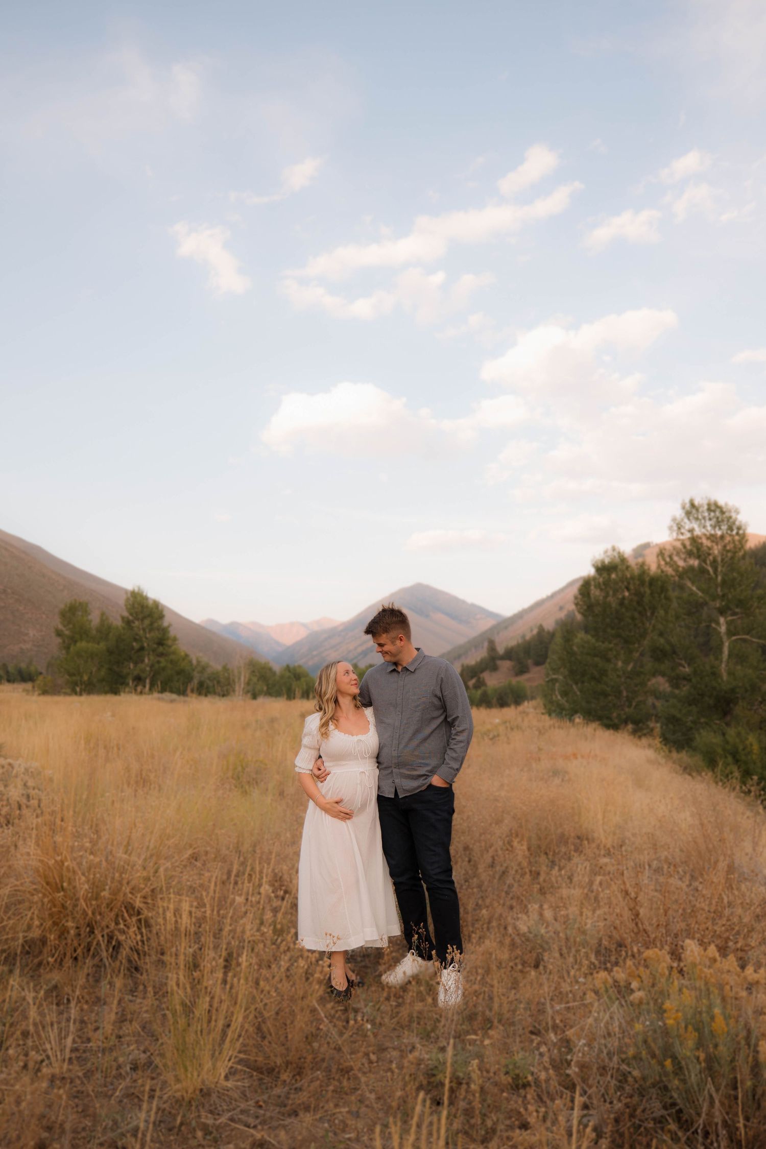 A couple stands together in a golden field with mountains in the background at sunset.