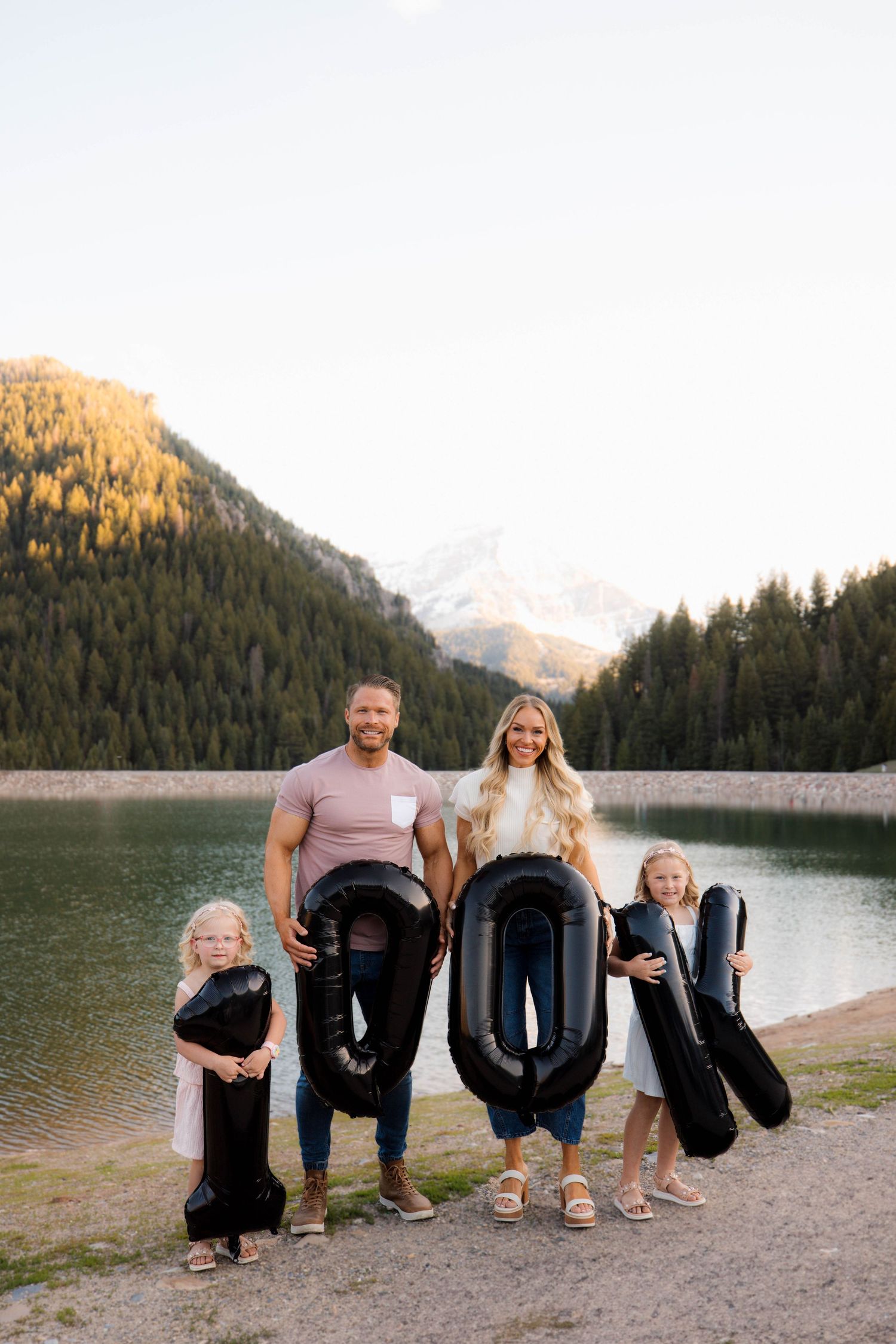 Family holds large black number balloon letters spelling 1000 while standing at a scenic mountain lake at sunset.
