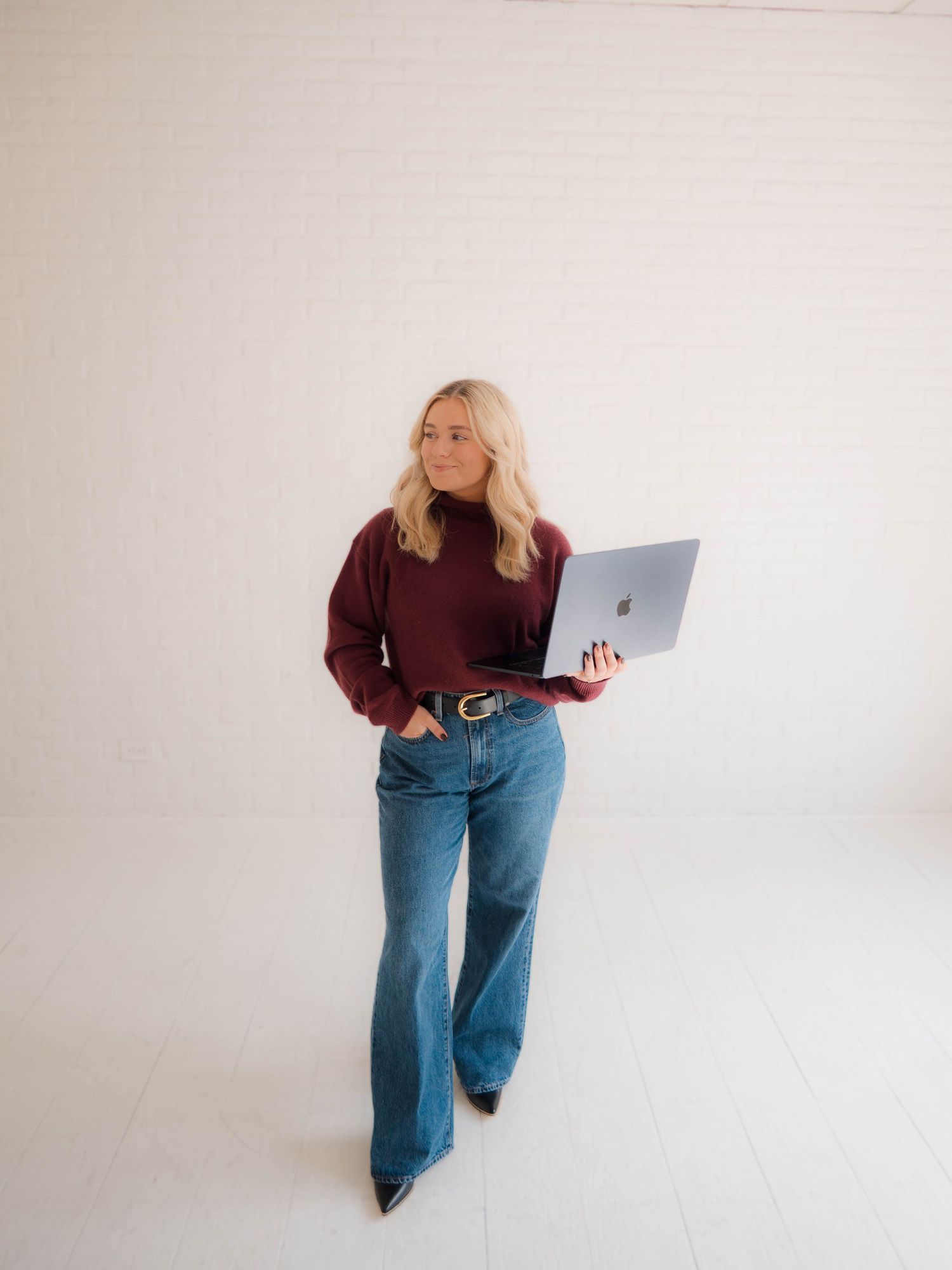 A person in a burgundy sweater and wide-leg jeans poses with a laptop against a white wall in four different positions.