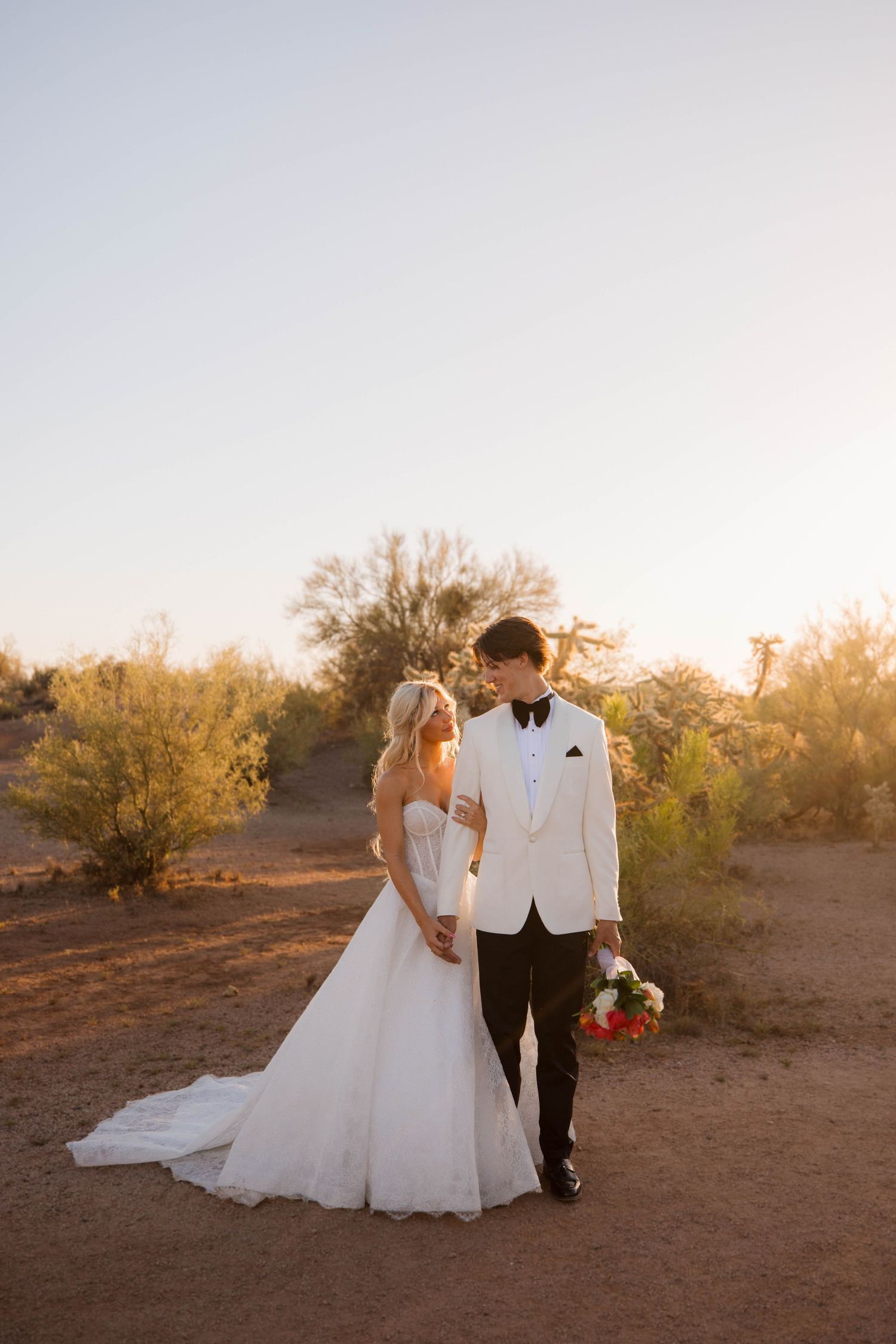 A couple in wedding attire stands together in a desert setting during golden hour sunset.