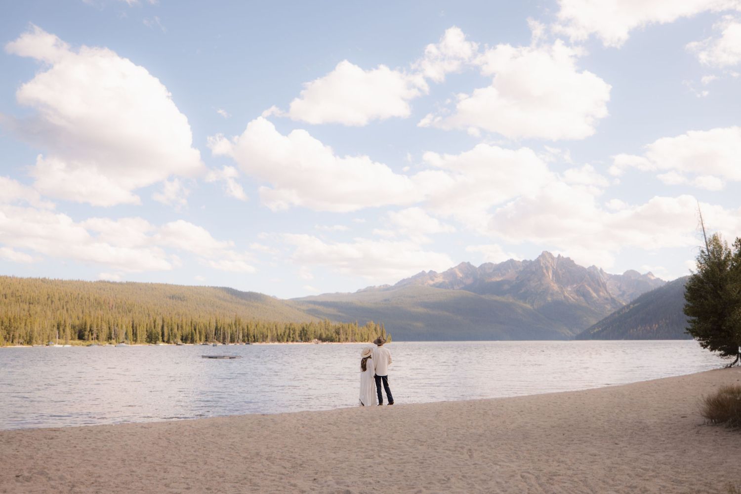 A figure stands on a peaceful beach shoreline at Redfish Lake with mountains and forest in the distance under blue skies.