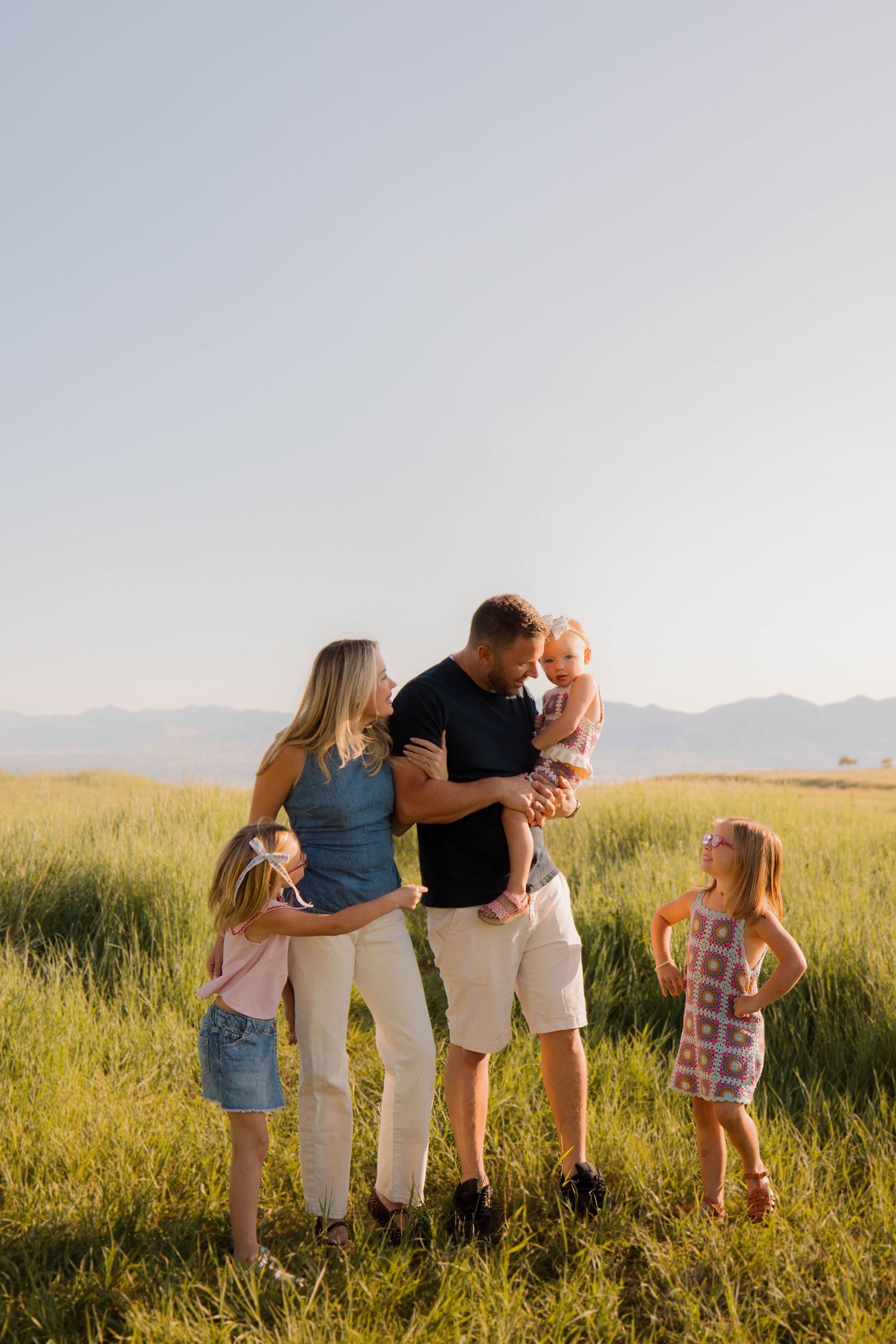 Family embracing in golden sunlit field with mountains in background during sunset golden hour.