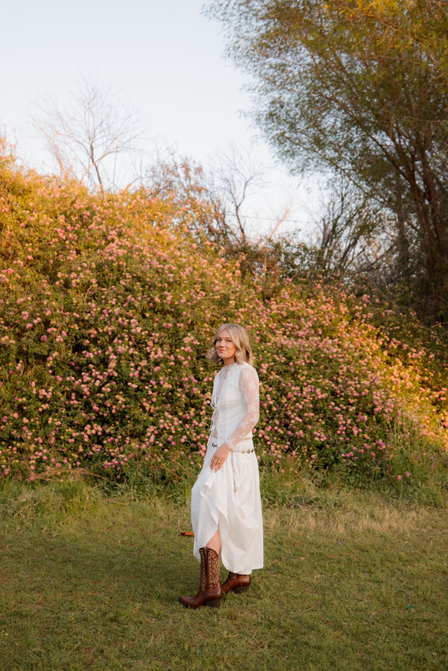A person in a white flowing dress twirls in a garden with autumn foliage and pink flowers in the background.