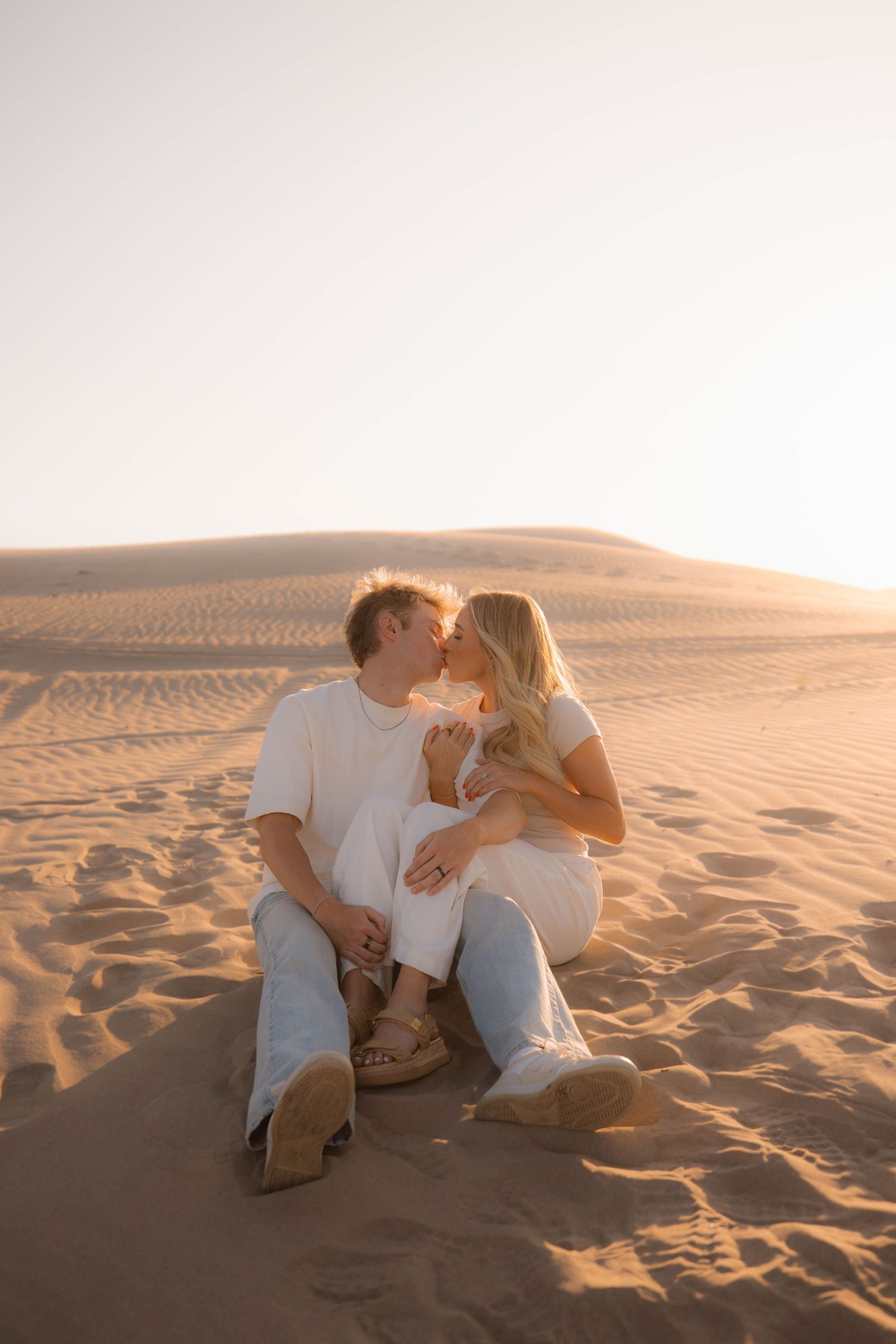 A romantic couple in white outfits share tender moments on sand dunes at sunset.