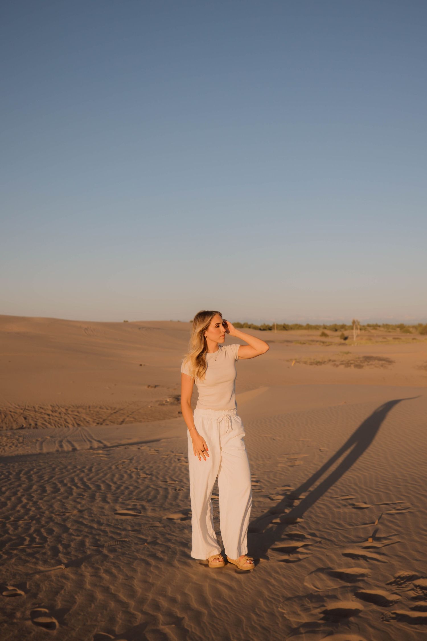 A person in white clothing walks across rippling sand dunes during golden hour in a desert landscape.