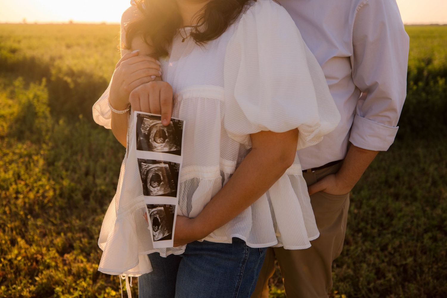 A couple holding a sonogram photo embraces in a golden sunlit field at sunset.
