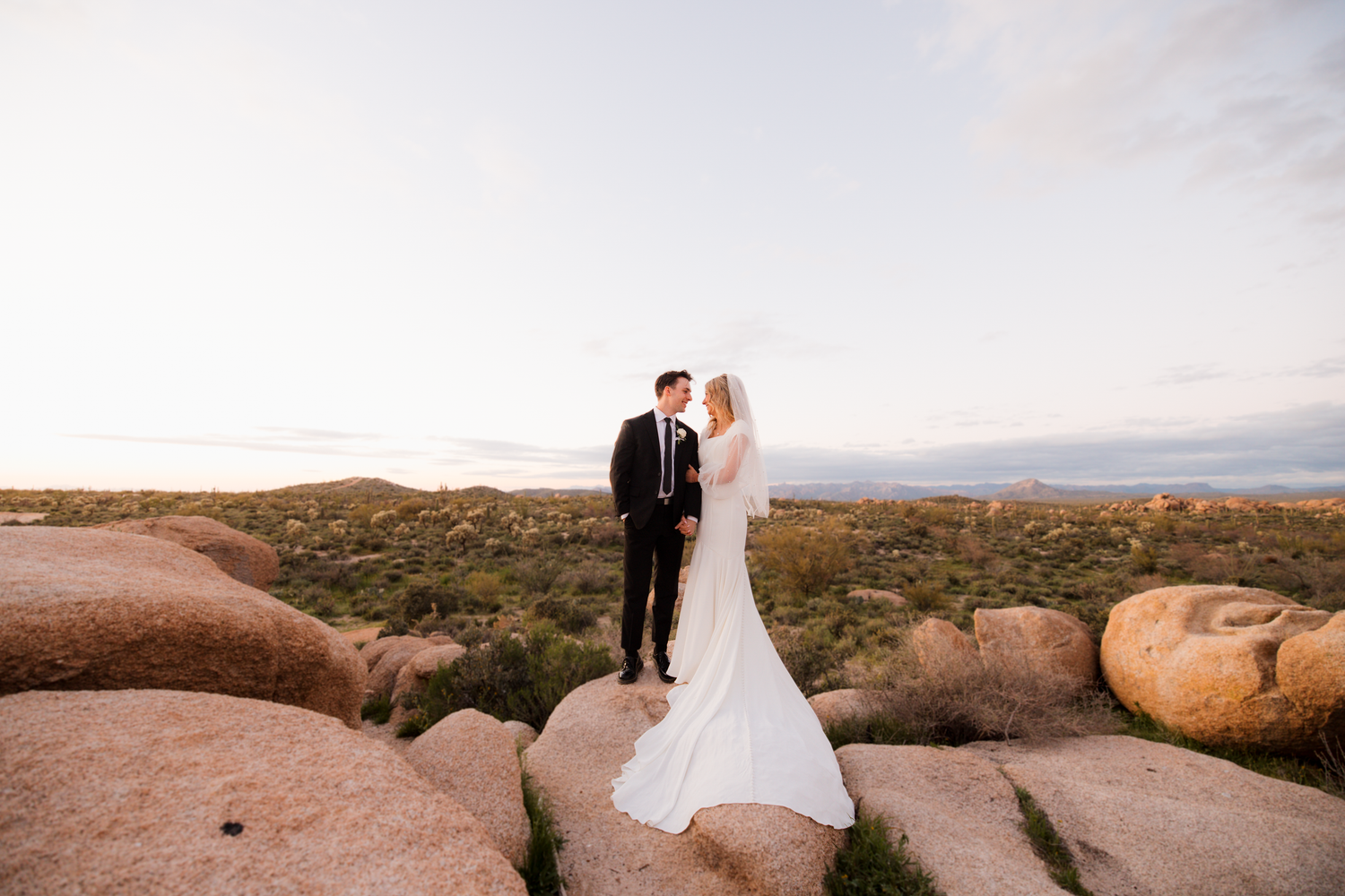 A couple in formal wedding attire stands atop red desert rocks during sunset in a dramatic desert landscape.
