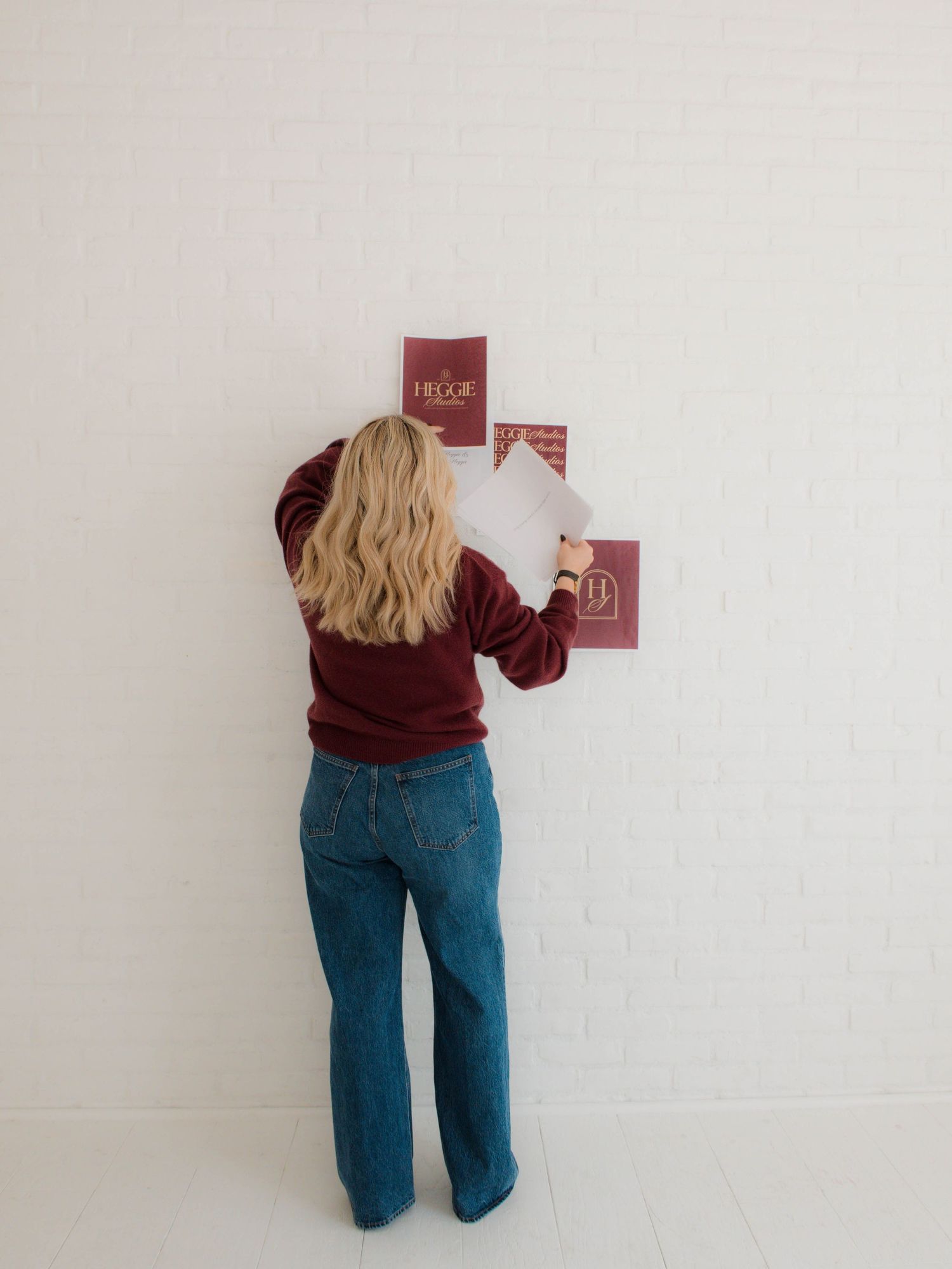 Someone in a burgundy sweater and blue jeans stands against a white wall holding small red cards.