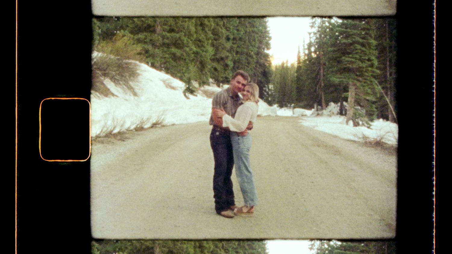 A couple embraces on a snow-covered path surrounded by evergreen trees during golden hour lighting.