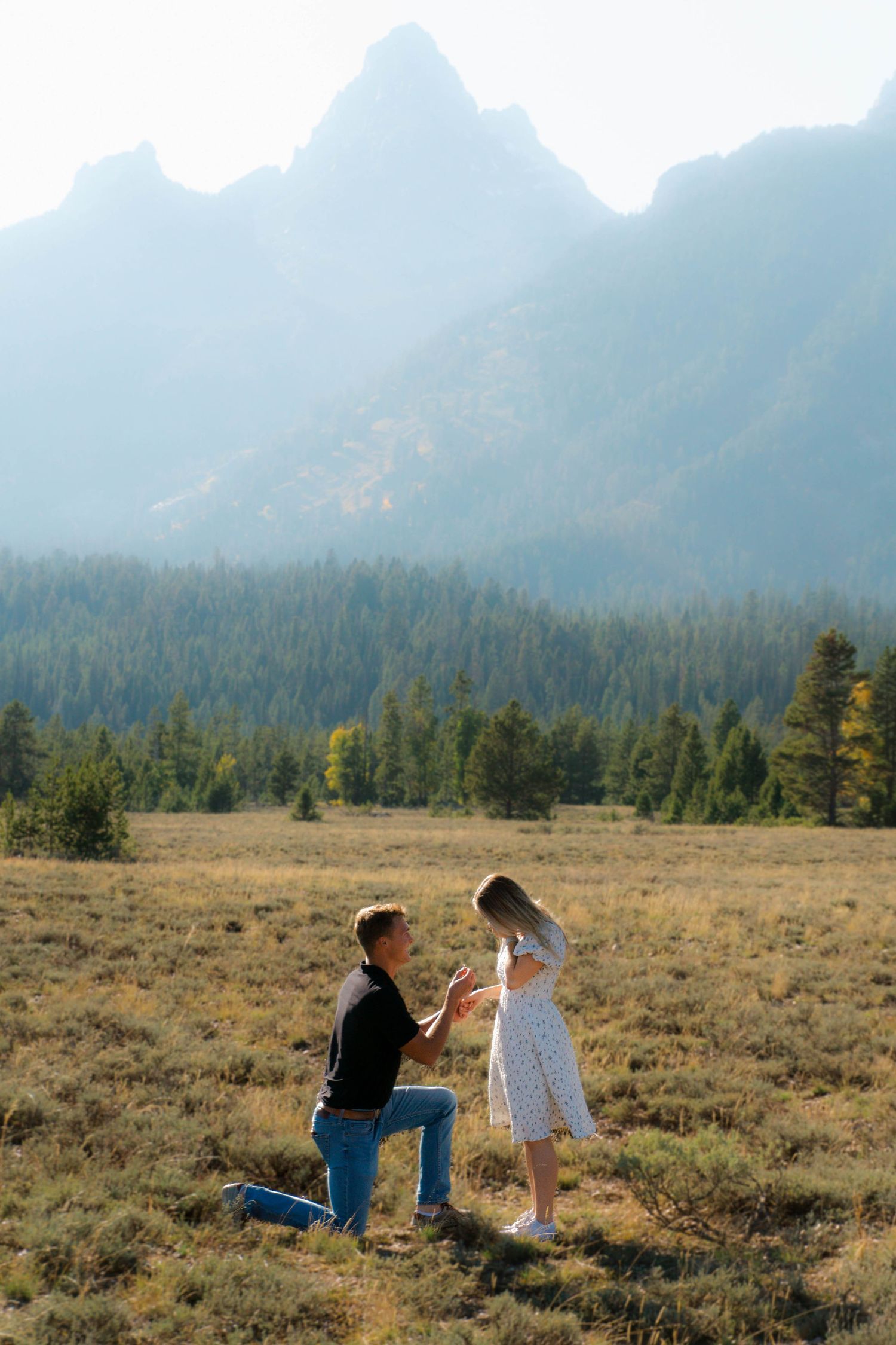 Two people sit on a grassy field with majestic mountain peaks and evergreen forest in the misty background.