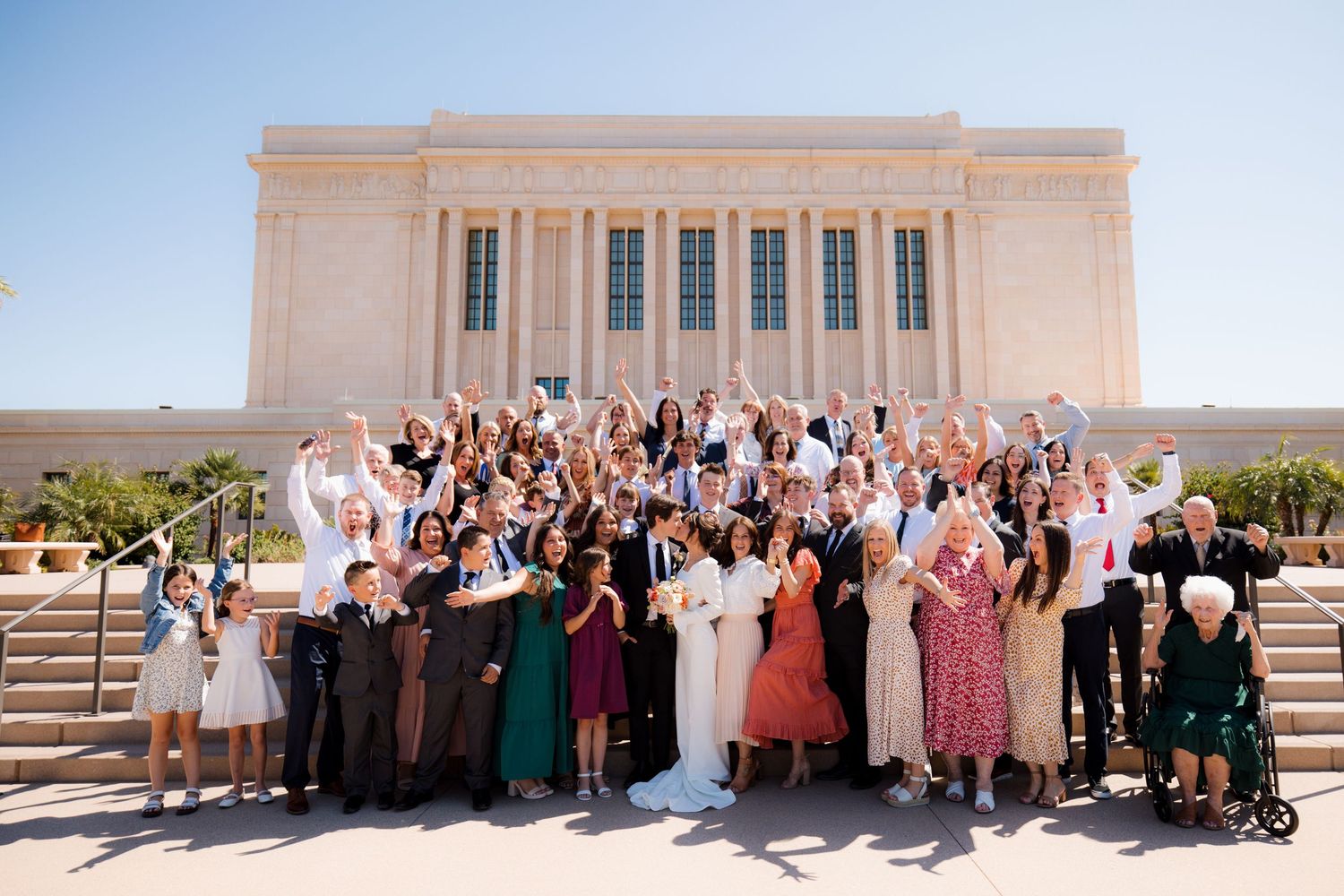 A large celebratory group photo on the steps of a light-colored temple building with classical architectural features.