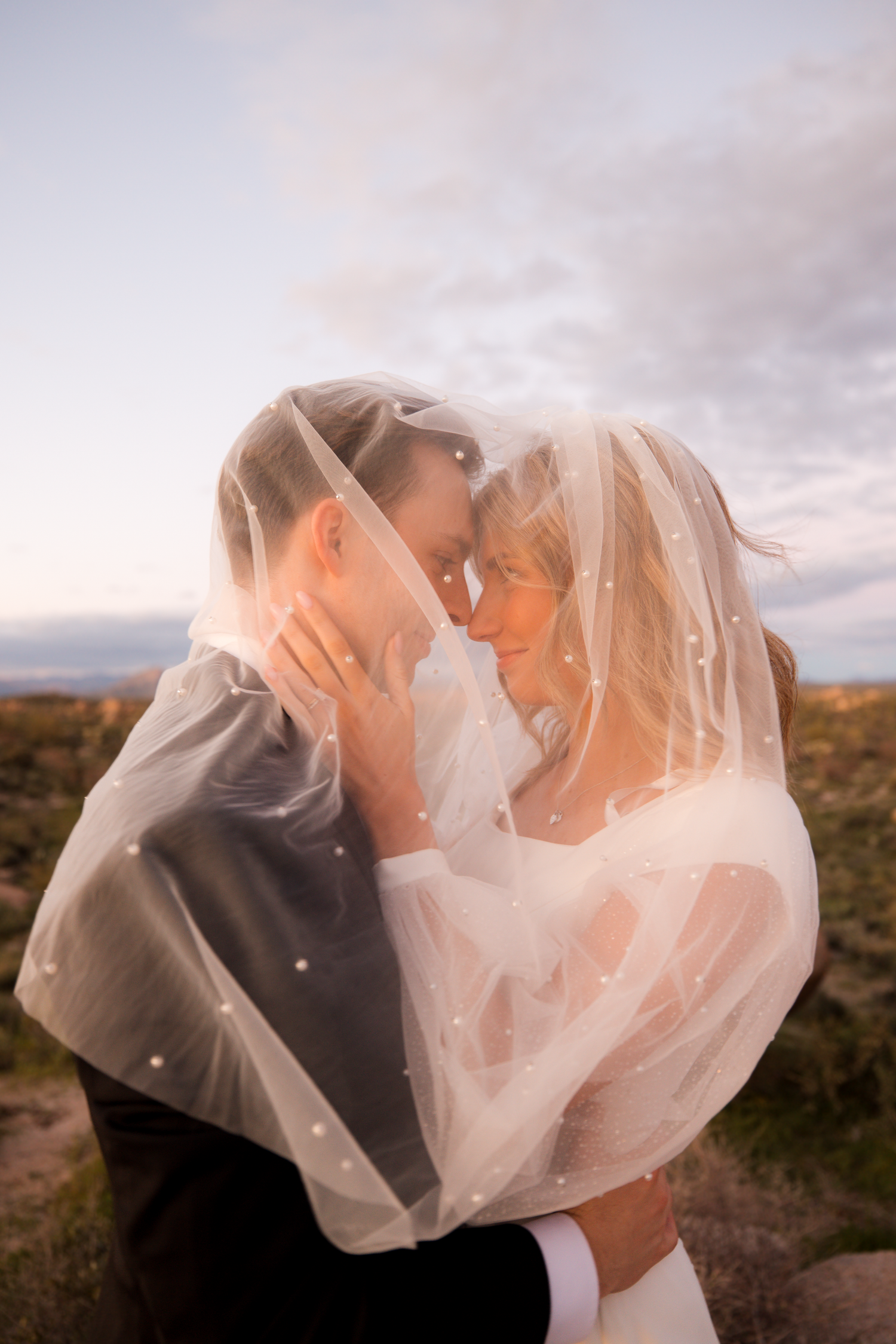 Romantic wedding couple shares intimate kisses under a flowing veil against a desert landscape at sunset.