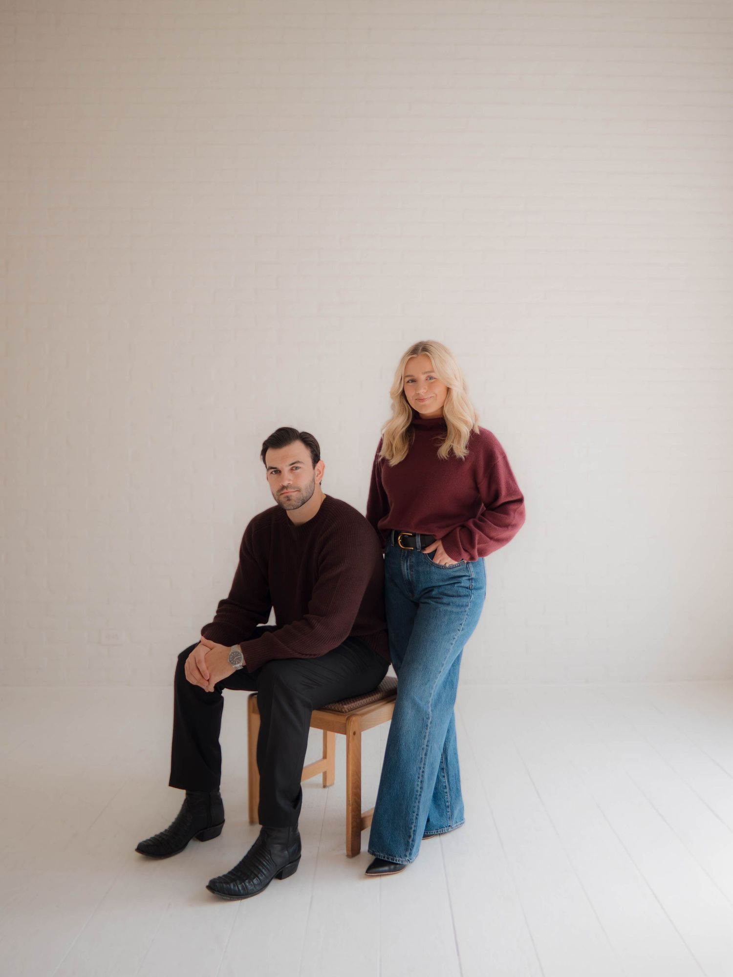 Couple in casual attire poses together in a minimalist studio setting with a beige background.